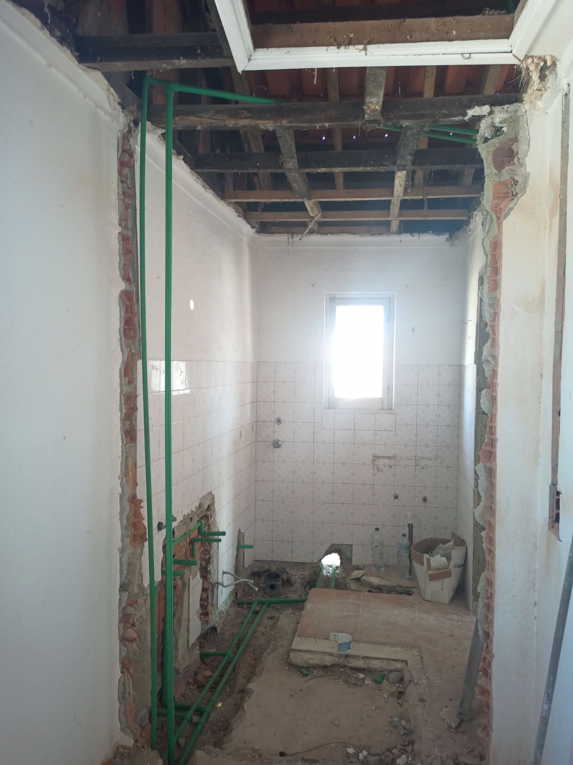 Banheiro em reforma, com encanamento exposto, paredes danificadas, vigas visíveis no teto e uma janela pequena.