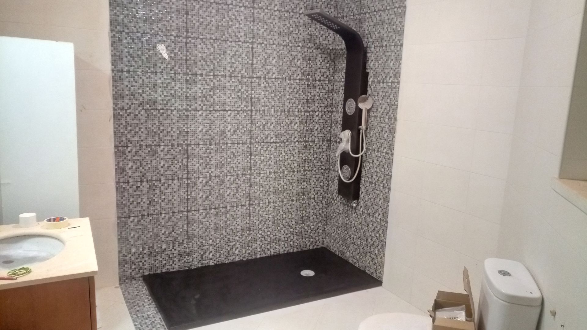 Banheiro com box de chuveiro revestido com azulejos pretos e cinzas, chuveiro preto e pia.
