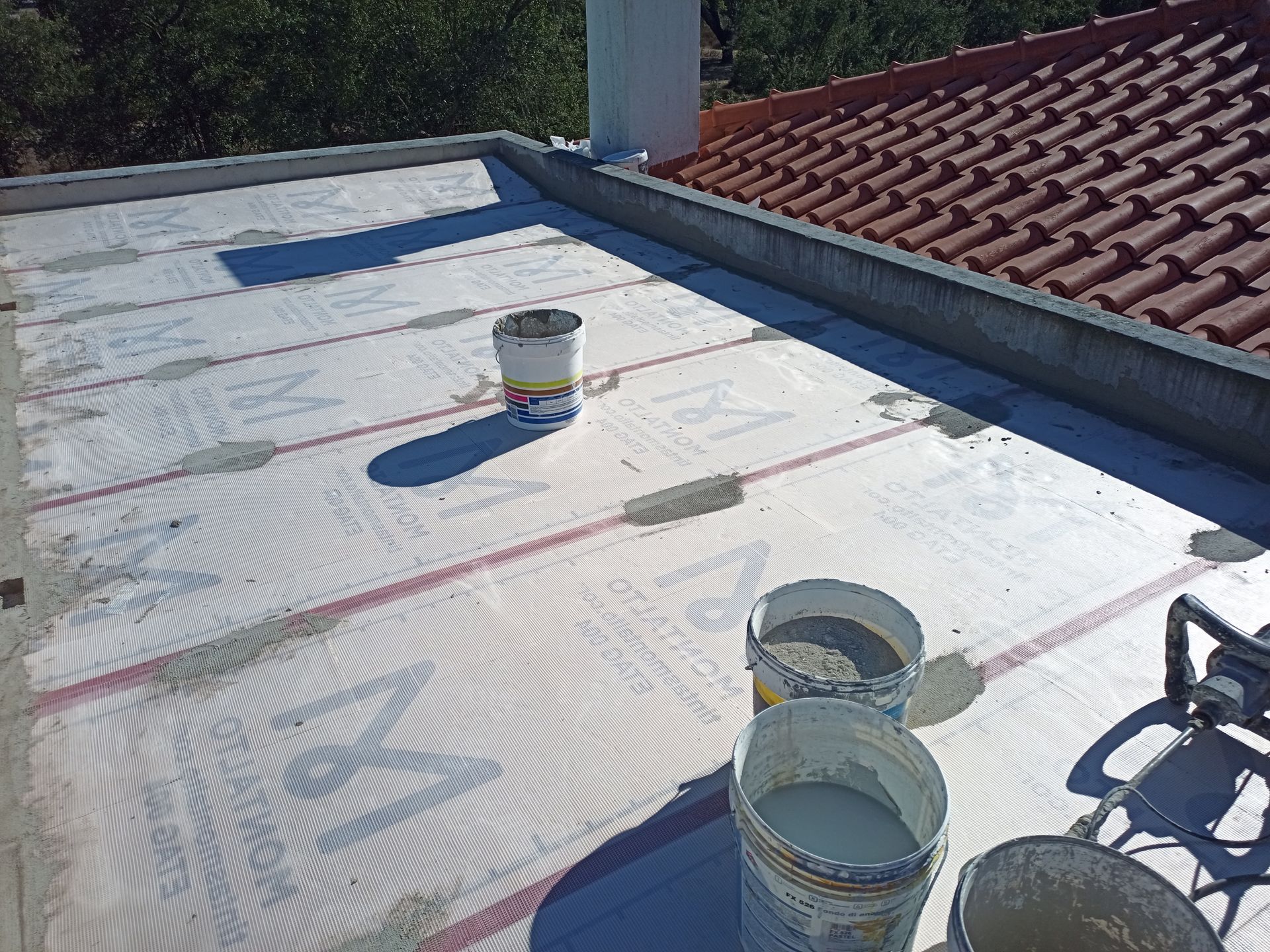 Telhado plano com isolamento branco, linhas vermelhas e baldes de material. Um segundo telhado, de telhas, está ao fundo.