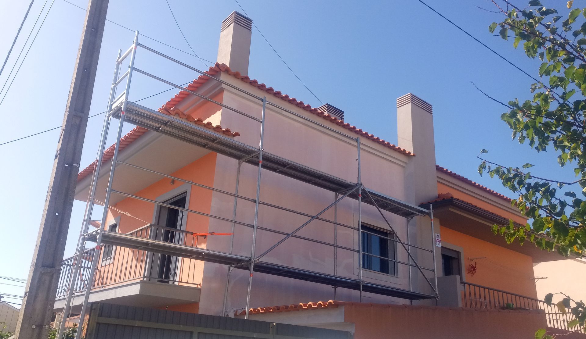 Exterior de uma casa com andaimes; paredes cor de pêssego, telhado vermelho, chaminés, céu azul.