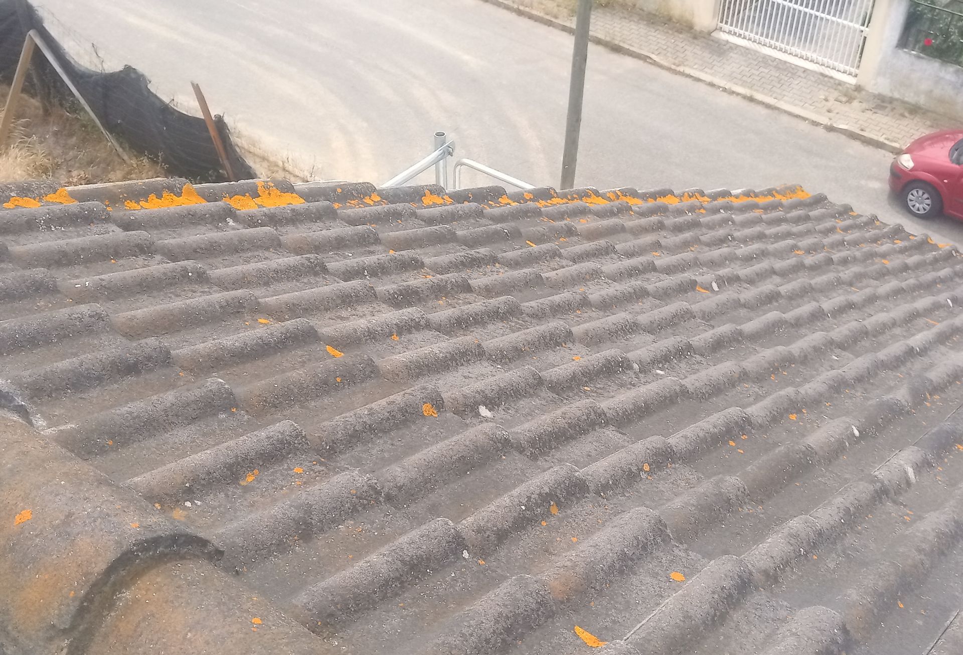 Telhado de telhas cinzentas com manchas de líquen alaranjado, com vista para uma rua e um carro vermelho estacionado.