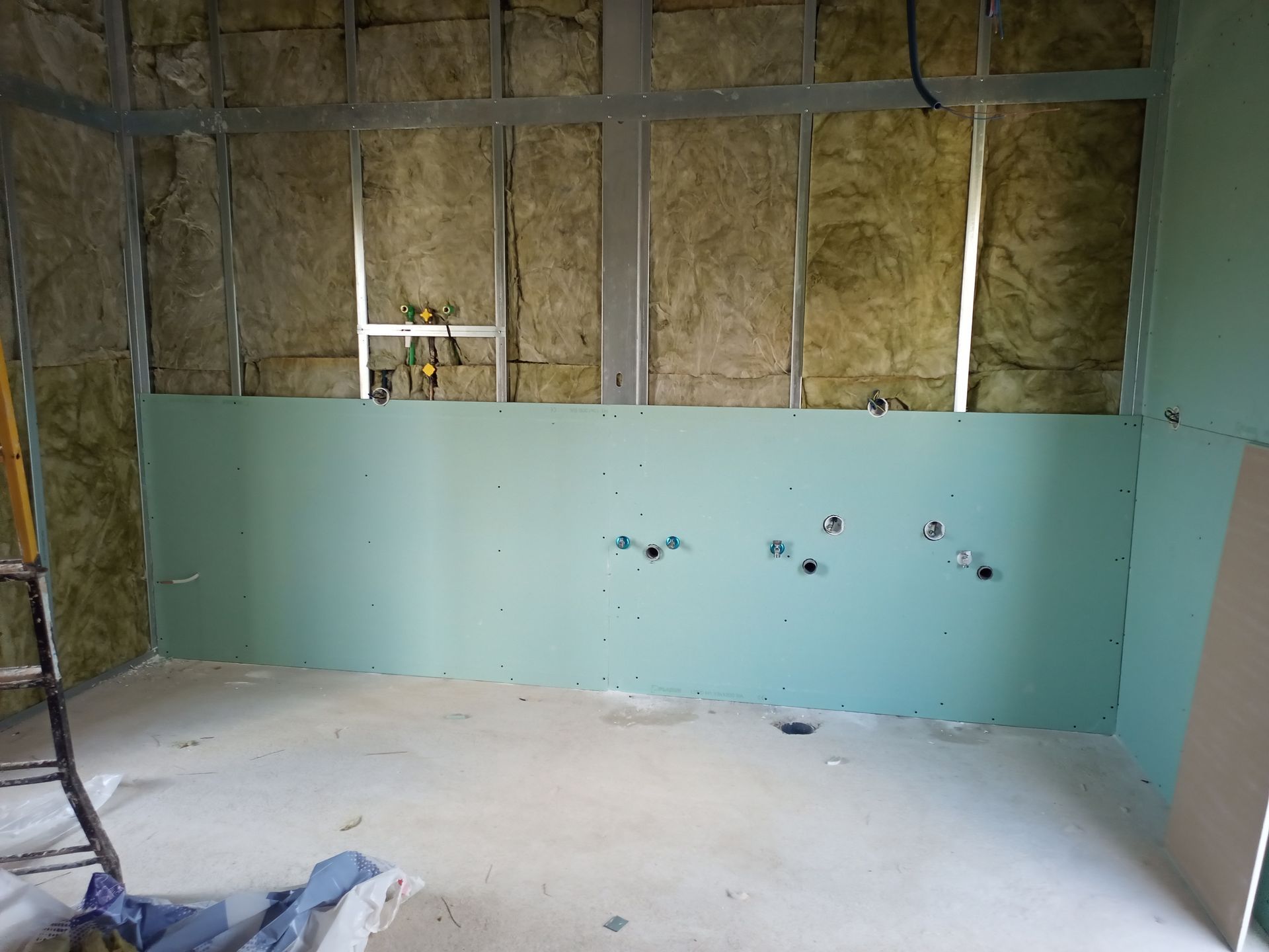 Canteiro de obras interno com drywall verde na parte inferior da parede, perfis metálicos, isolamento e encanamento exposto.