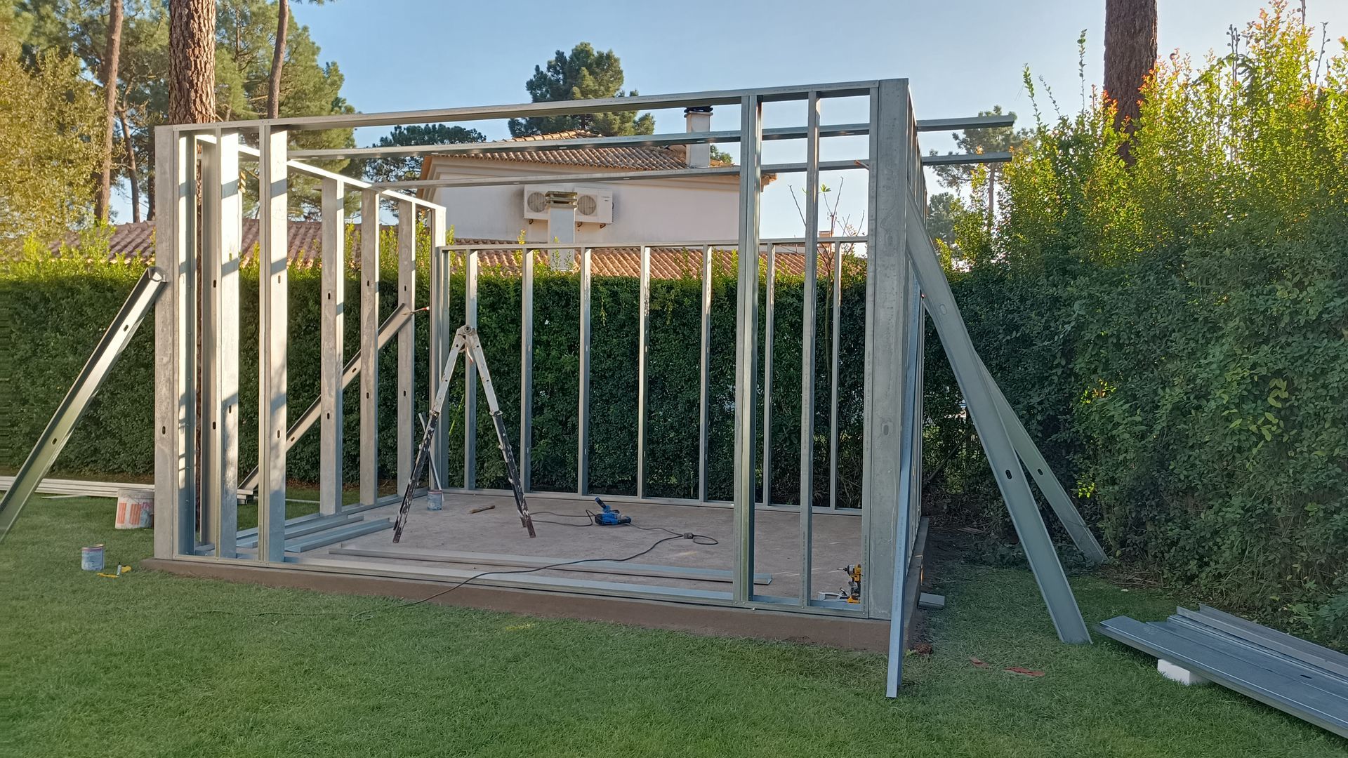 Estrutura metálica de um pequeno galpão em construção, situada na grama, com vegetação ao fundo.