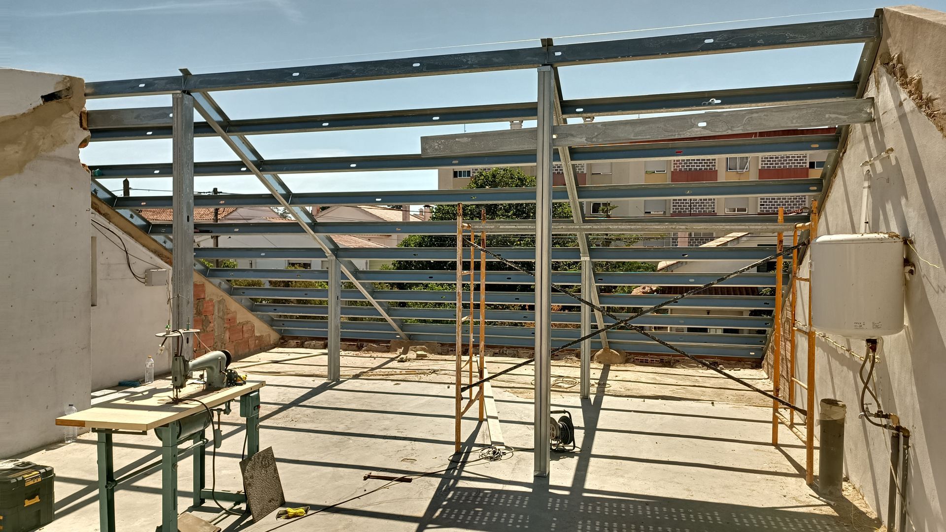 Estrutura metálica em um telhado, possivelmente para construção. Andaimes, bancada de trabalho e edifícios visíveis ao fundo.