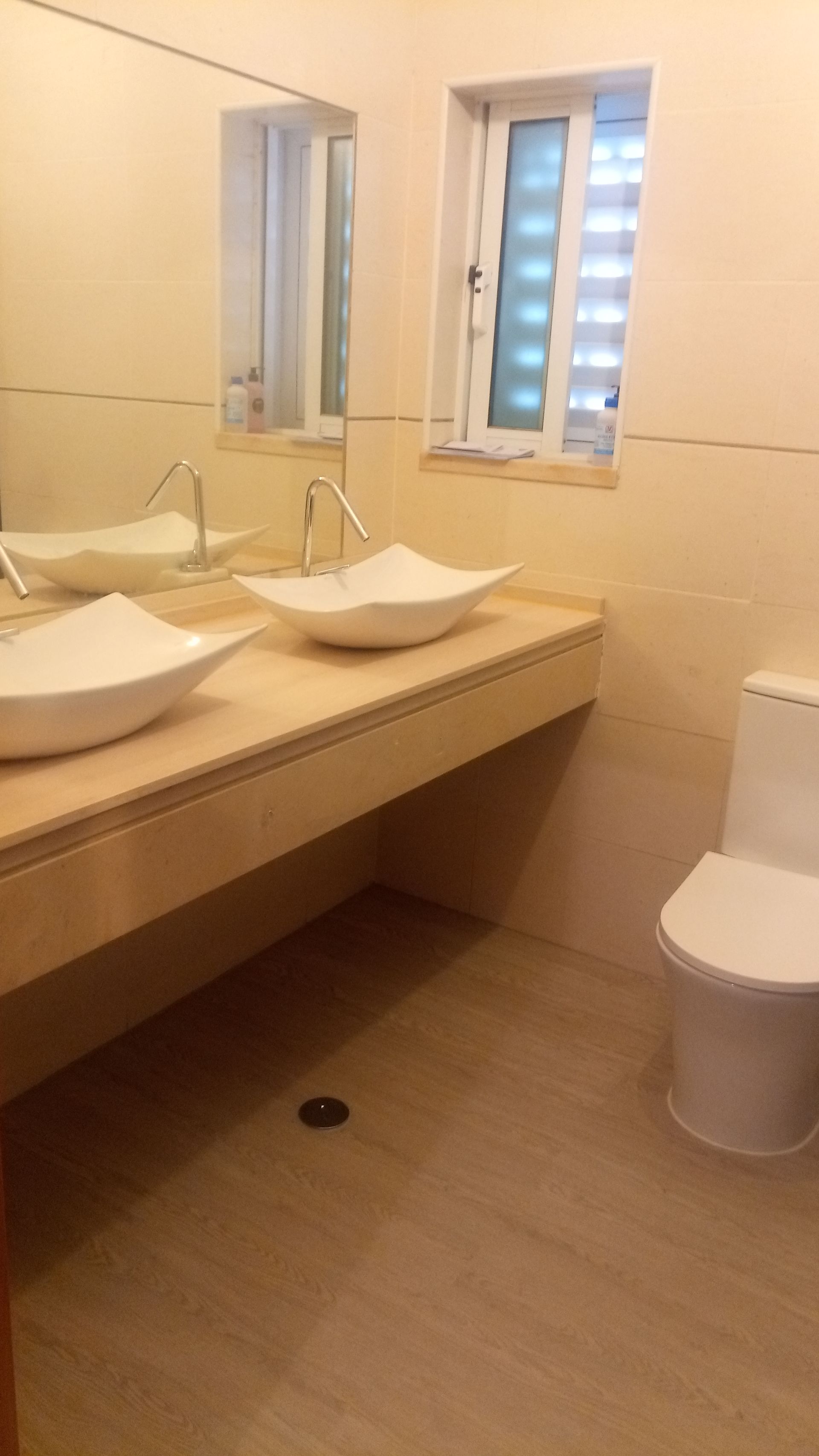 Banheiro com duas pias brancas sobre uma bancada de cor clara, espelho grande e vaso sanitário.