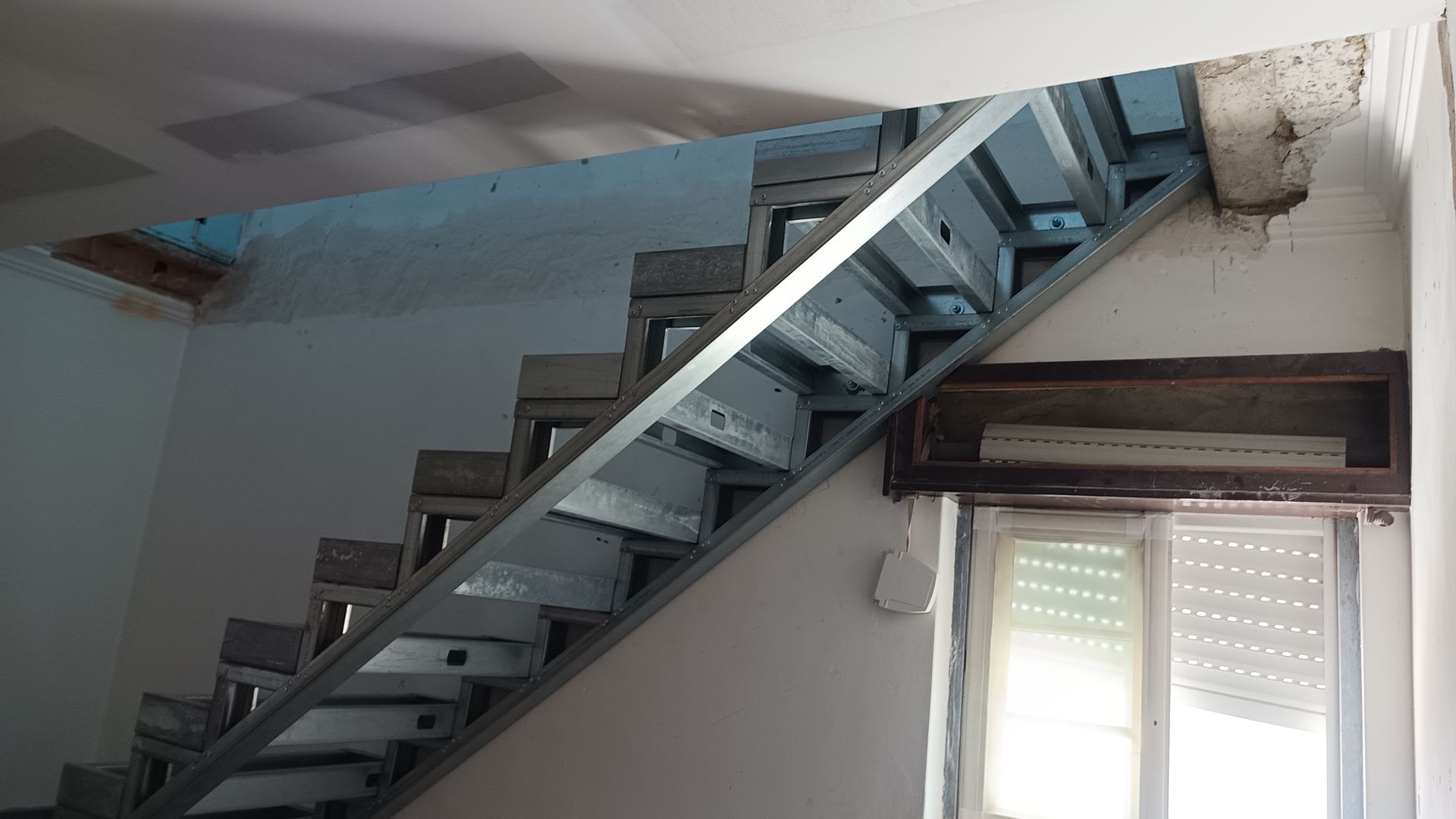 Escada metálica em construção, emoldurada contra uma parede branca.