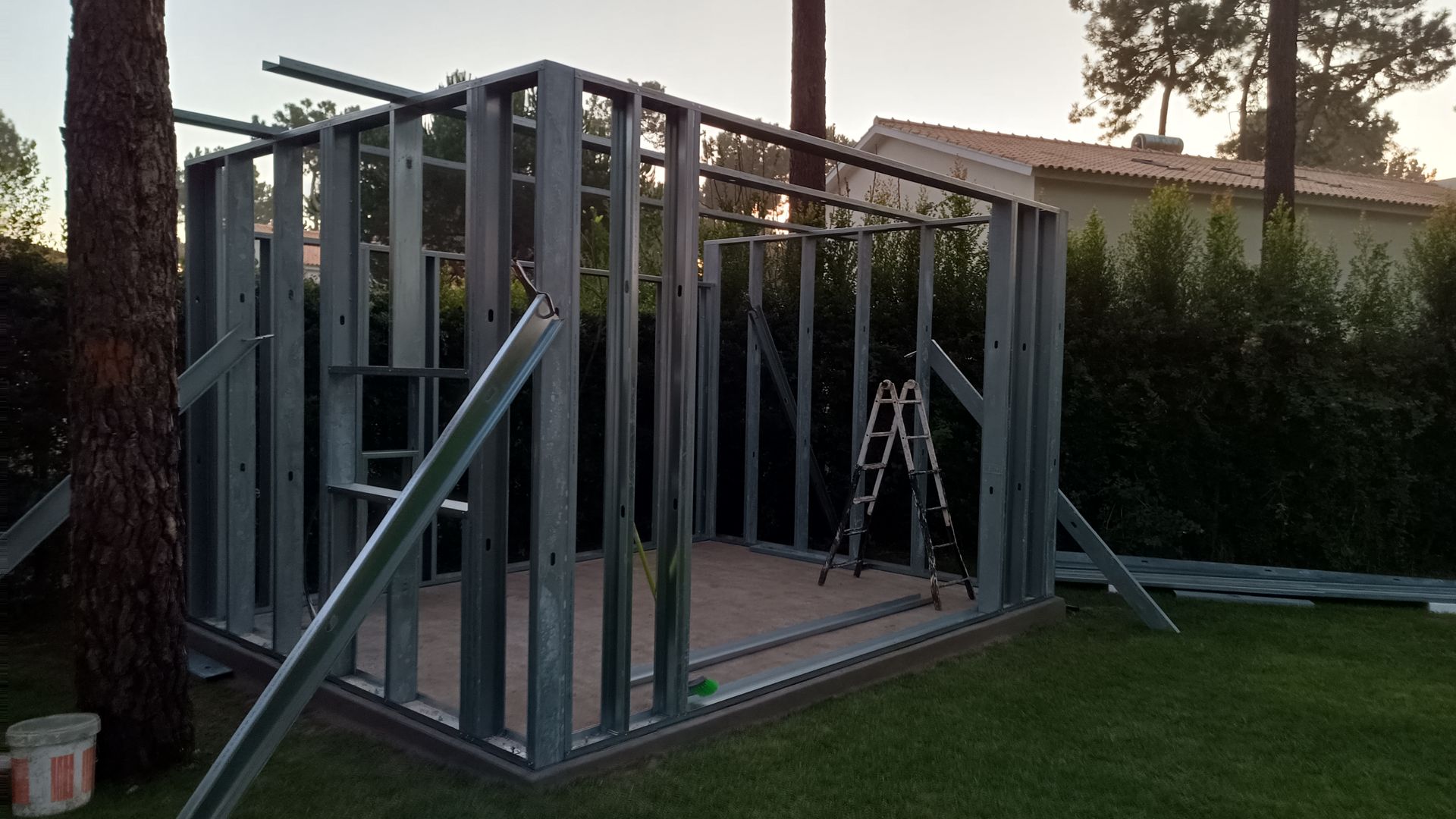 Estrutura metálica de um pequeno galpão em construção, situado em um quintal gramado com árvores.