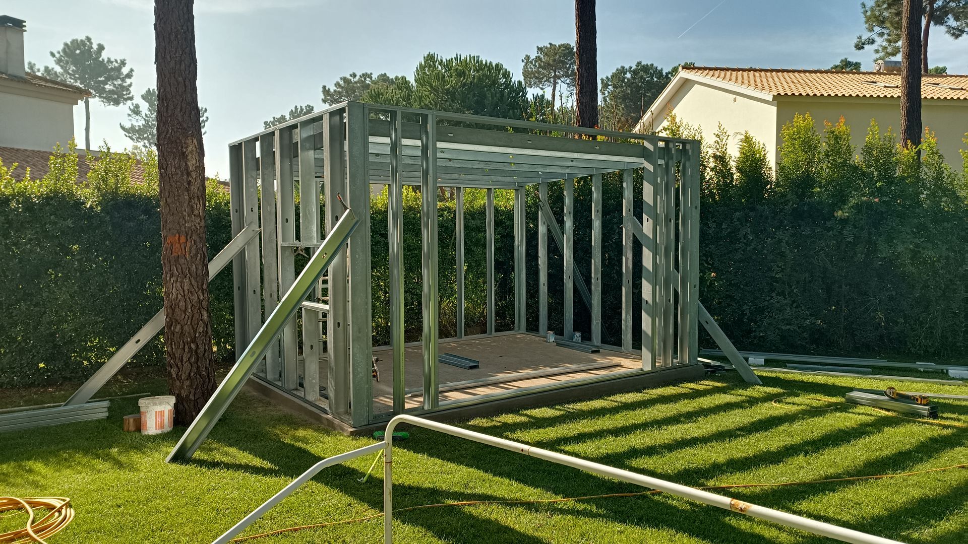 Estrutura metálica de uma pequena construção em andamento sobre grama verde, com árvores e uma casa ao fundo.