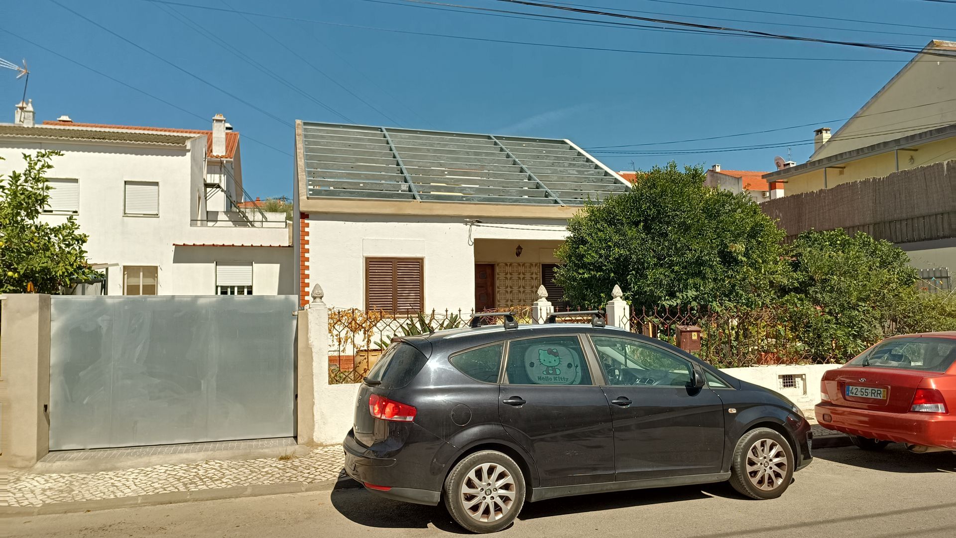 Uma pequena casa branca com telhado cinza, um carro escuro e um carro vermelho estacionados na rua. Céu azul.