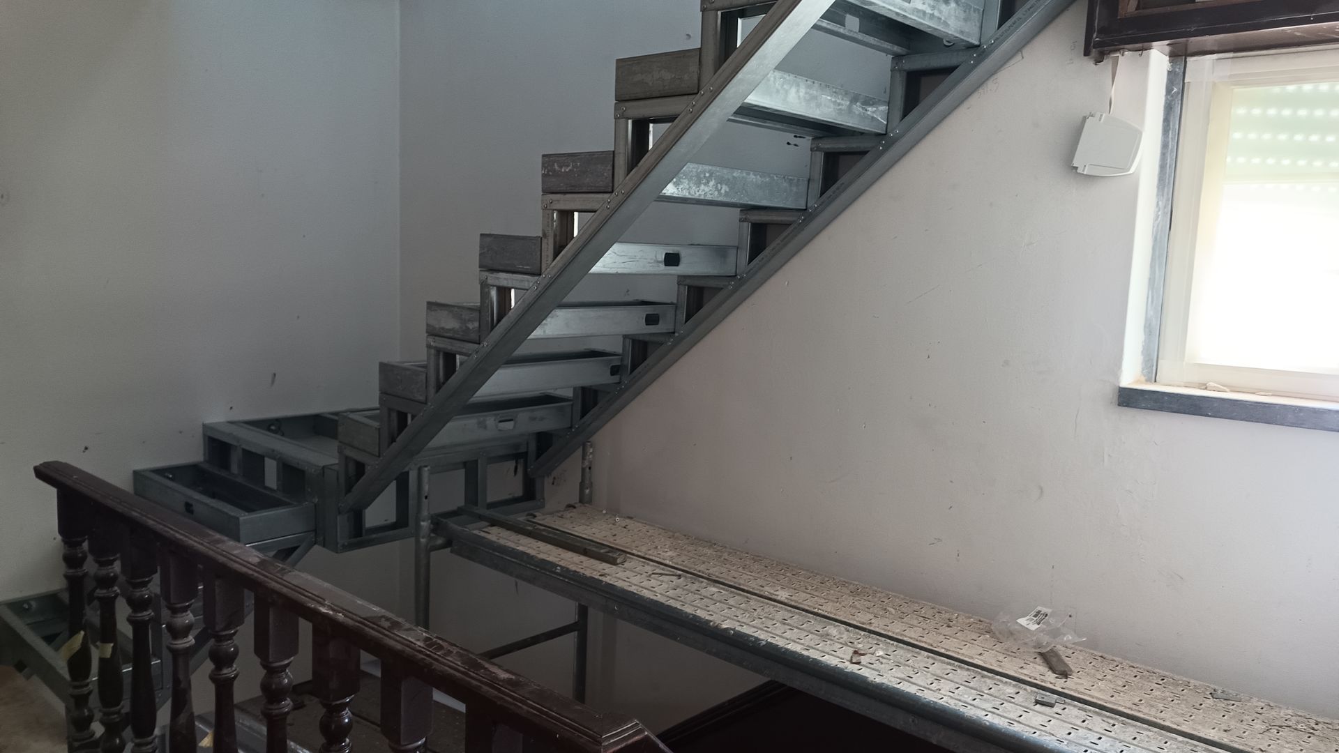 Escada metálica em construção, com degraus abertos, adjacente a uma parede e corrimão.