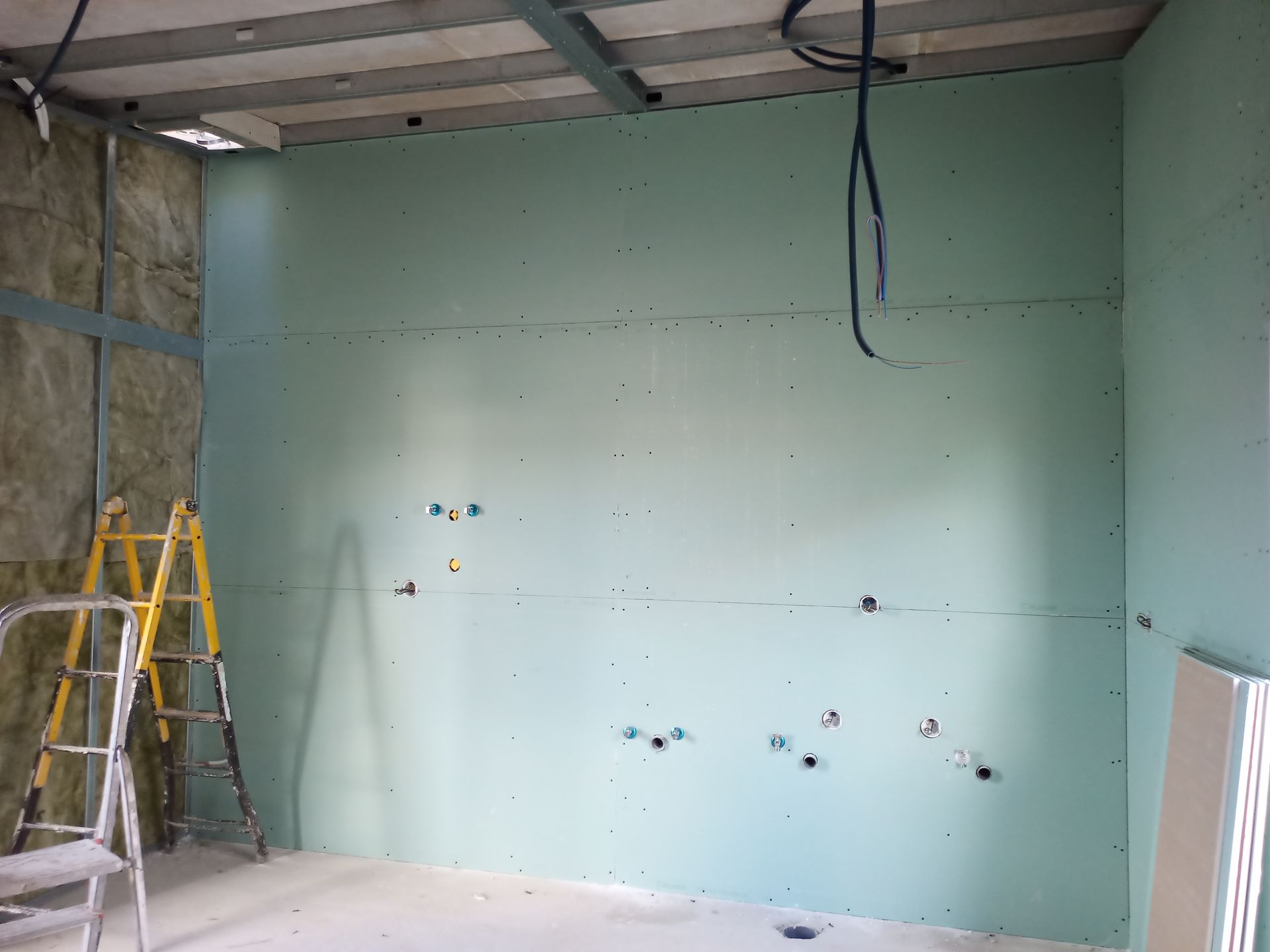 Construção interna: drywall verde em uma parede com furos para tomadas elétricas, escada e isolamento visíveis.