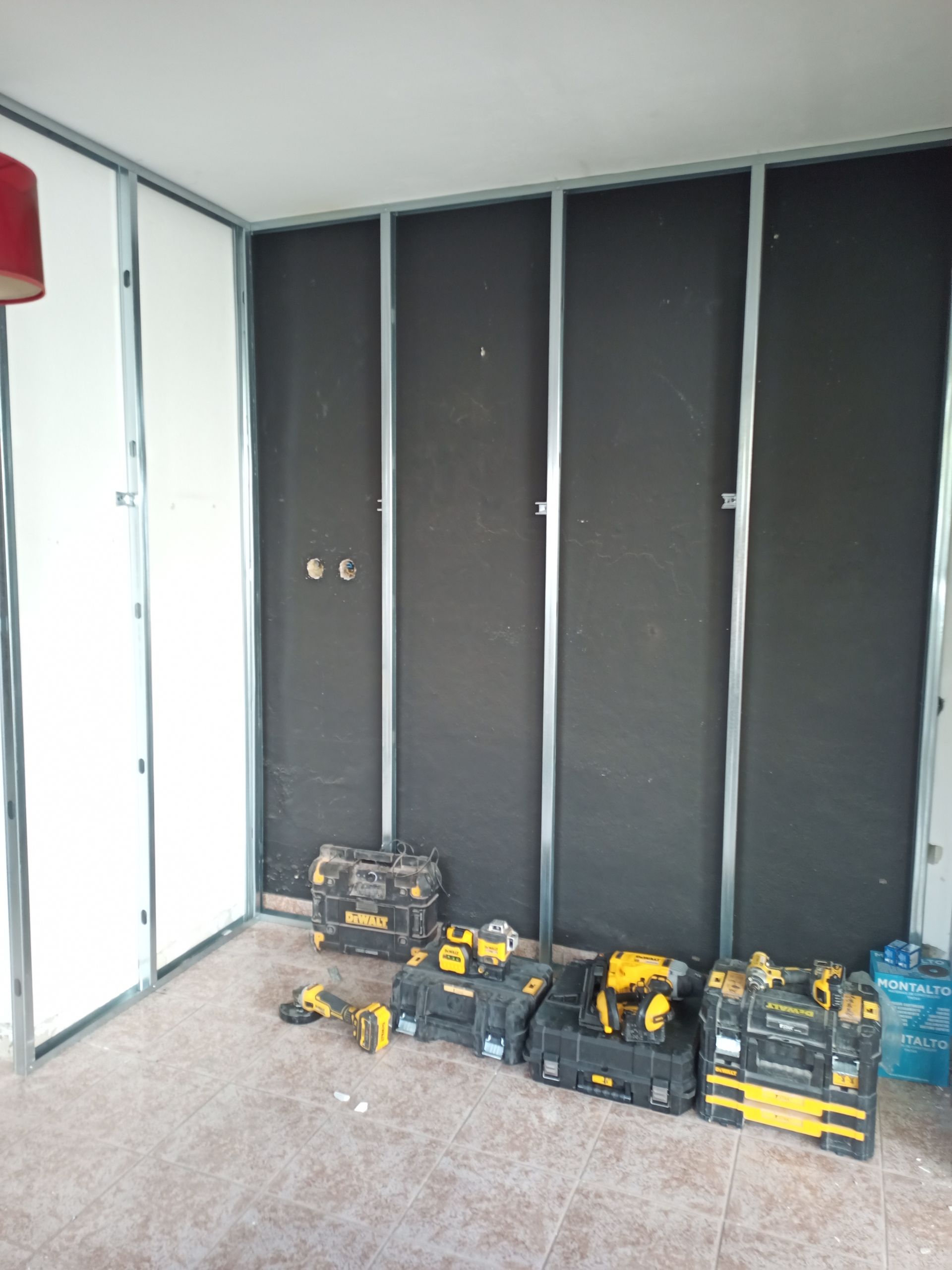 Interior de um cômodo em construção, com estrutura metálica, isolamento preto, painéis de parede brancos e ferramentas espalhadas.