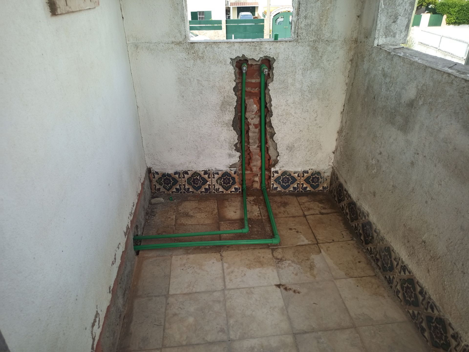 Tubulações de água verdes instaladas na parede do banheiro. Azulejos no piso e na base da parede. Paredes brancas e parcialmente danificadas.