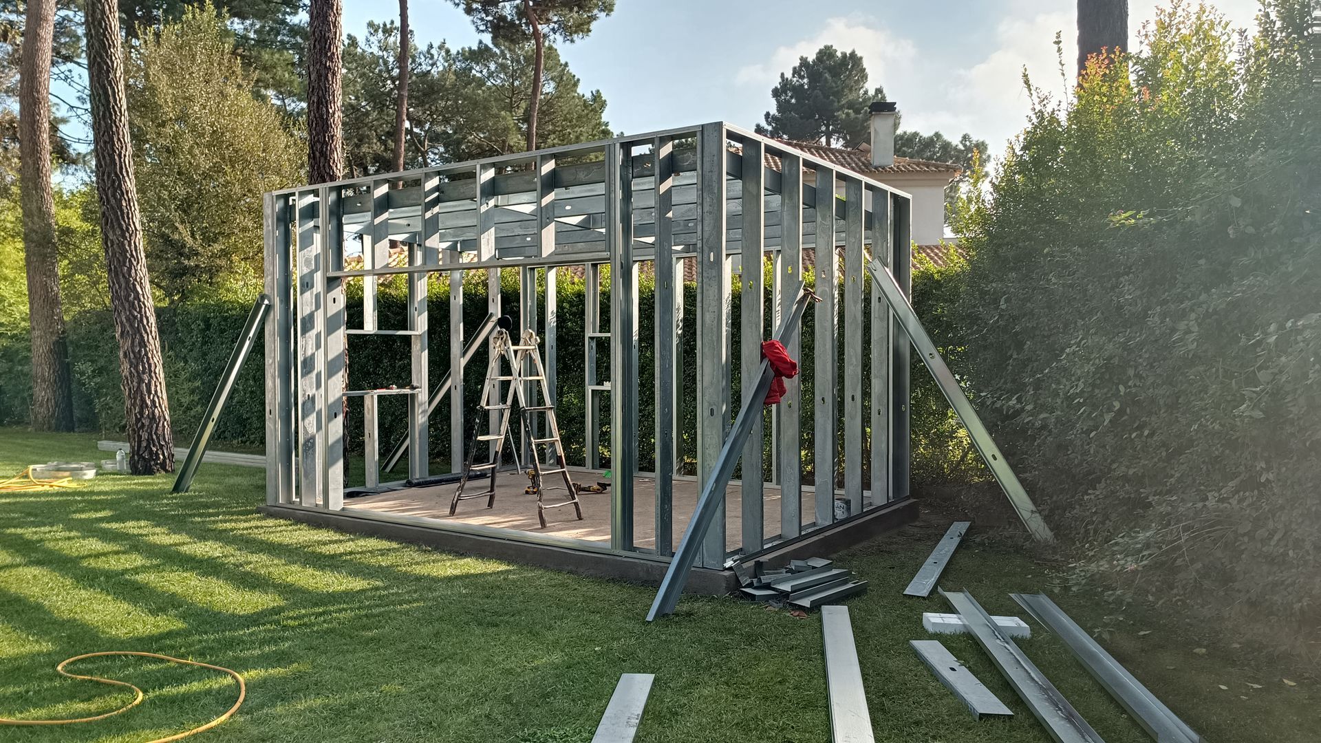 Estrutura metálica de um galpão em construção sobre um gramado; árvores e um prédio ao fundo.