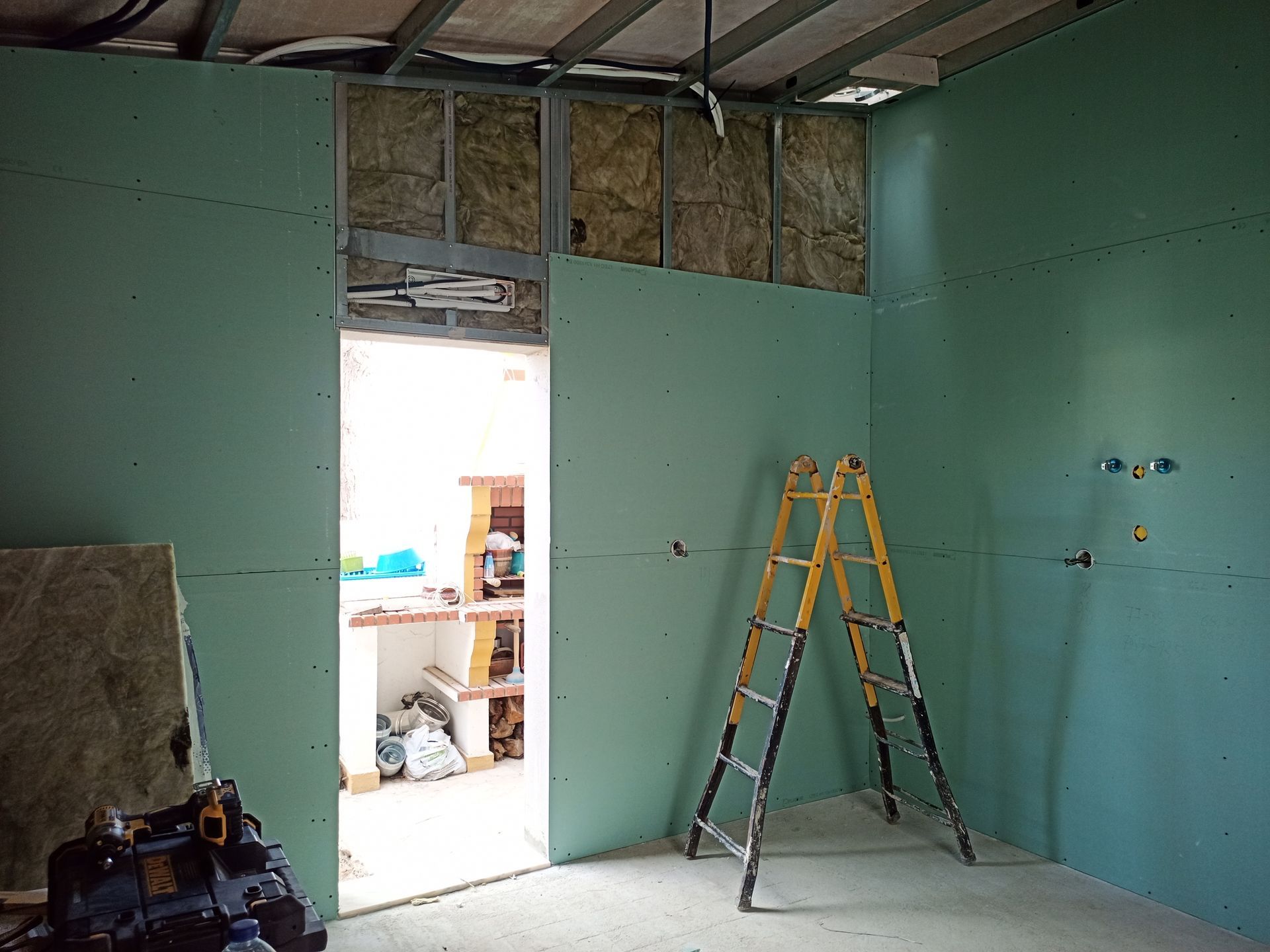 Interior de um cômodo em construção, paredes de gesso verde, isolamento exposto, porta aberta, escada amarela.
