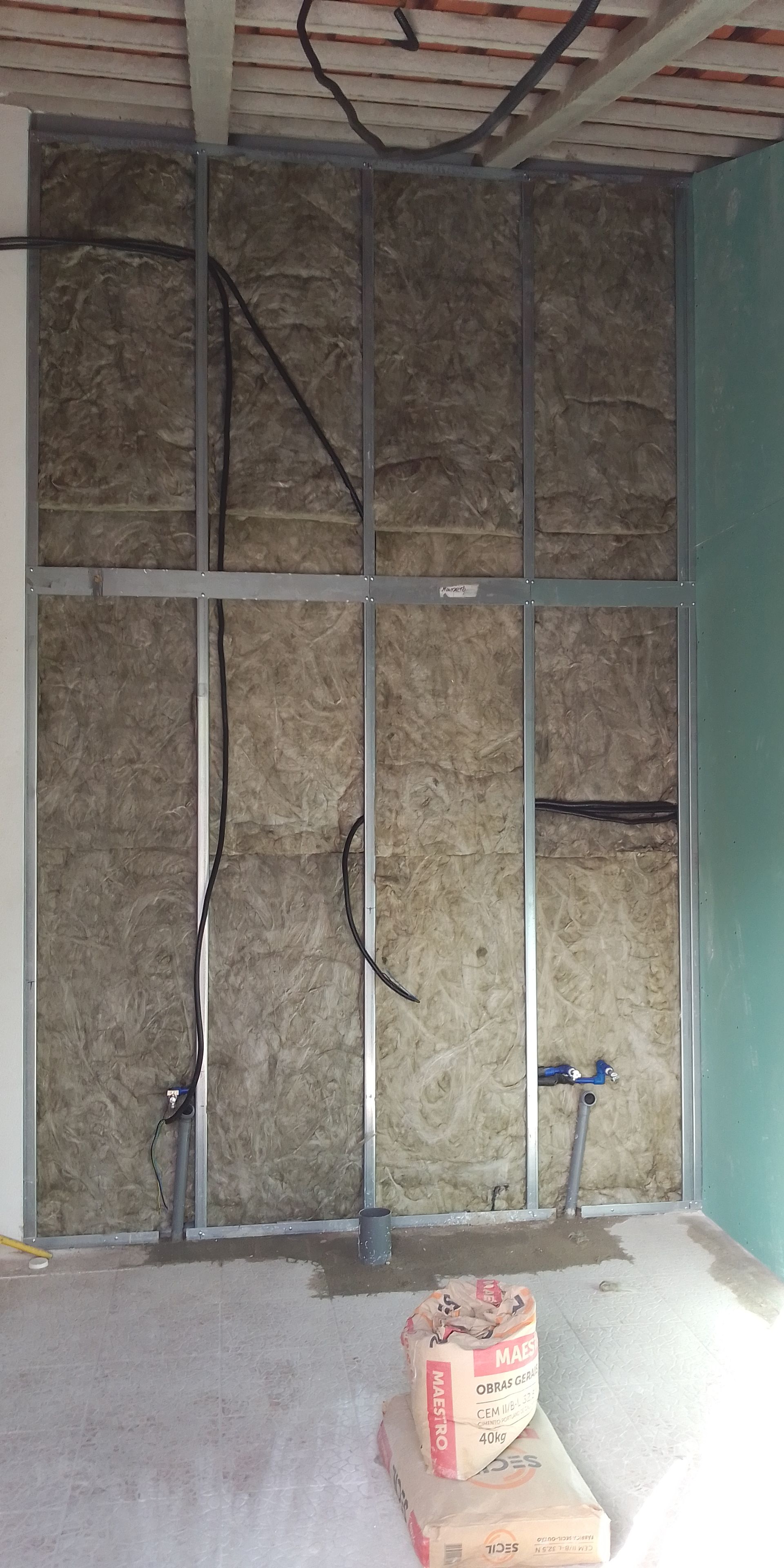 Construção de parede interna com estrutura metálica, isolamento e instalações elétricas e hidráulicas expostas. Um saco de cimento está no chão.