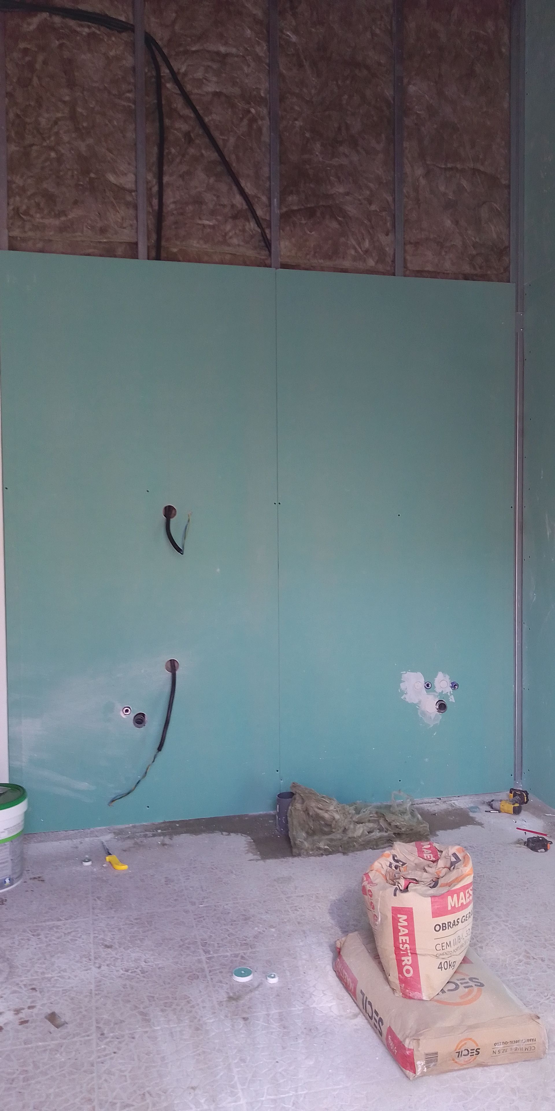 Interior de uma sala em construção com painéis de parede verdes e sacos de cimento no chão.