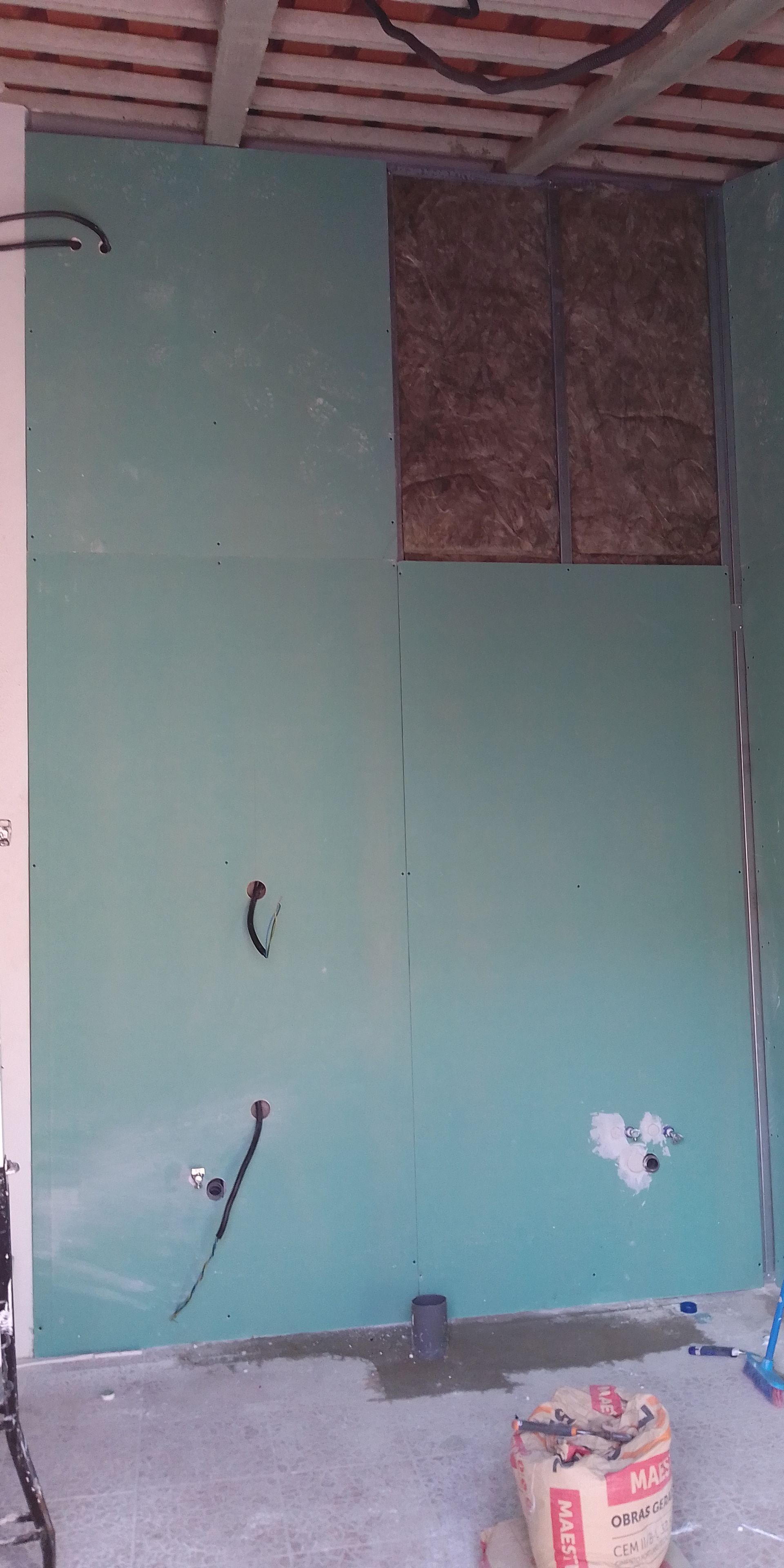 Placa de gesso verde instalada em uma parede com madeira exposta e um saco de material no chão.