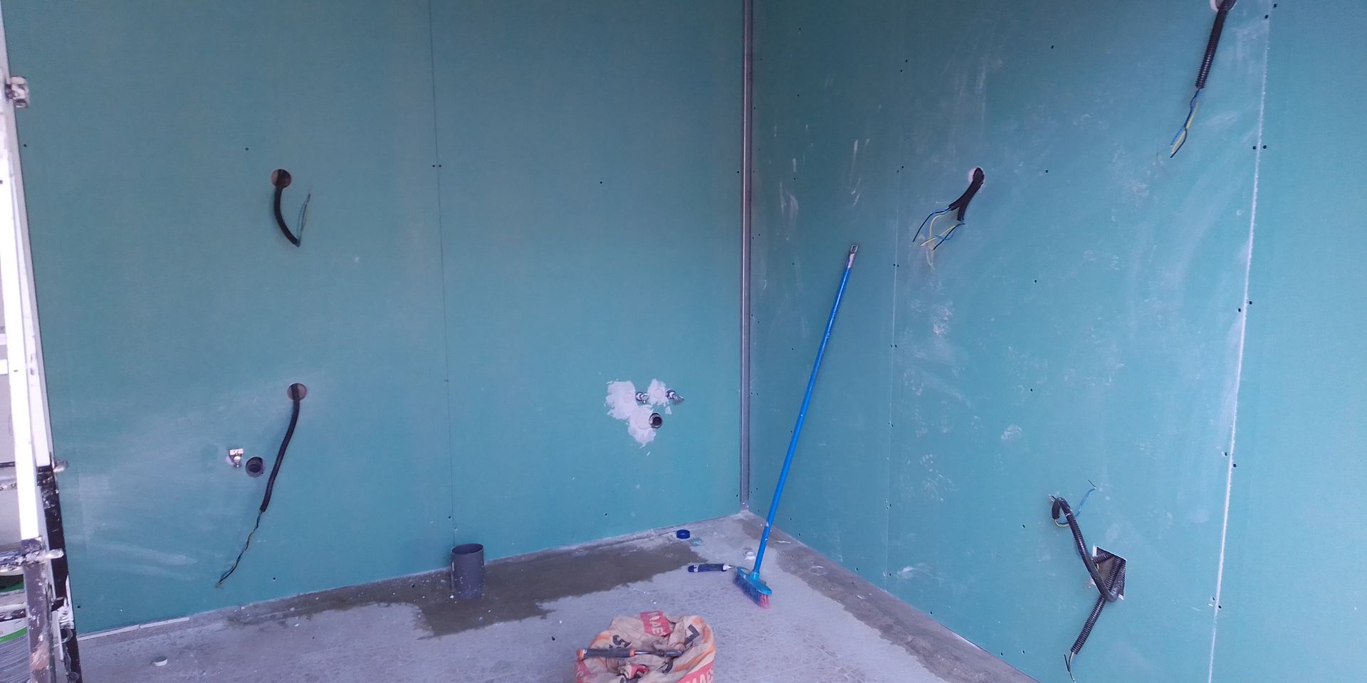 Parede interna com materiais e ferramentas de construção. Placa de gesso azul-esverdeada com selante preto, vassoura azul e piso de concreto.