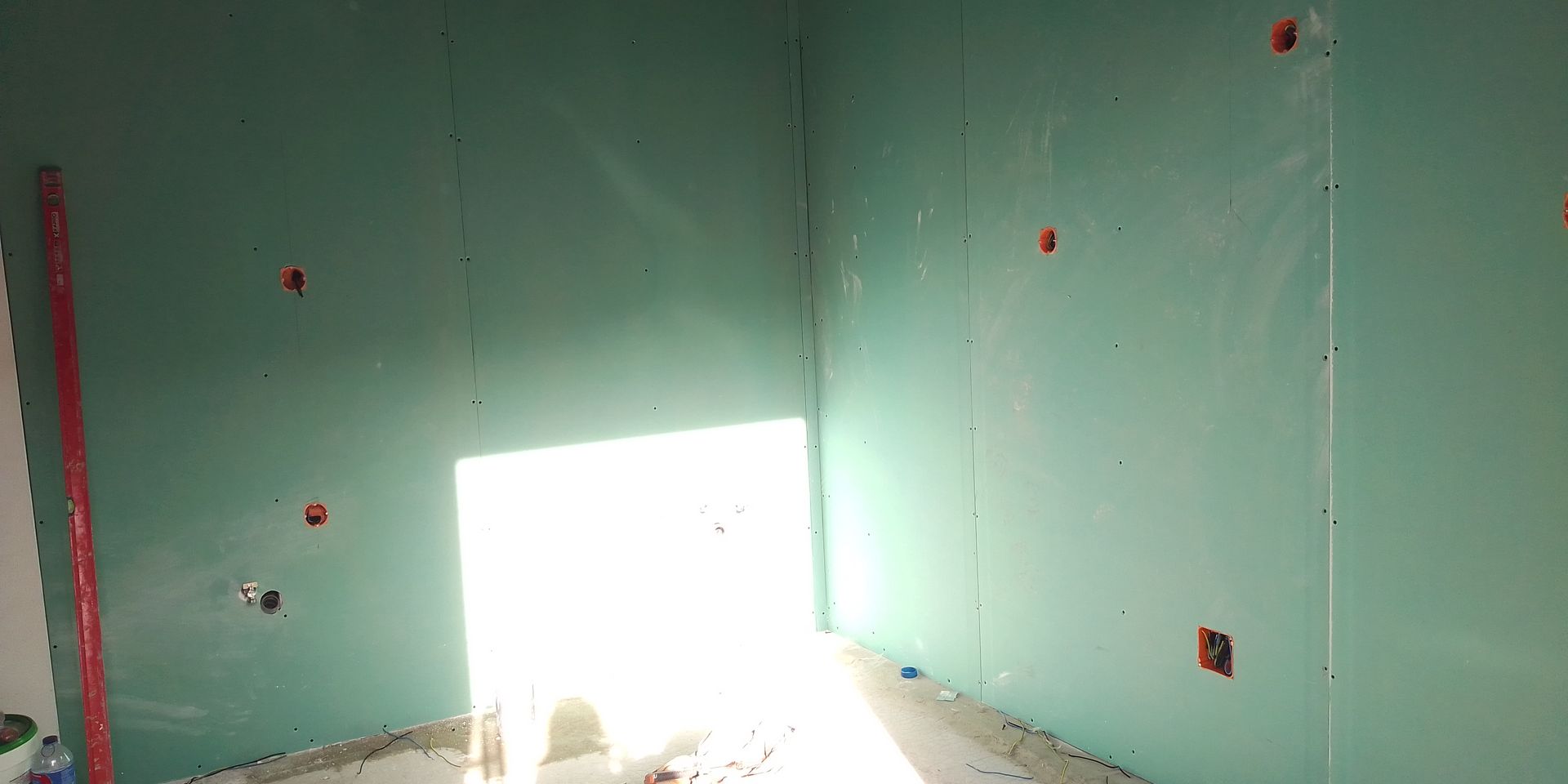 Vista interna de um cômodo com paredes revestidas de gesso acartonado. Uma luz forte entra pela porta.