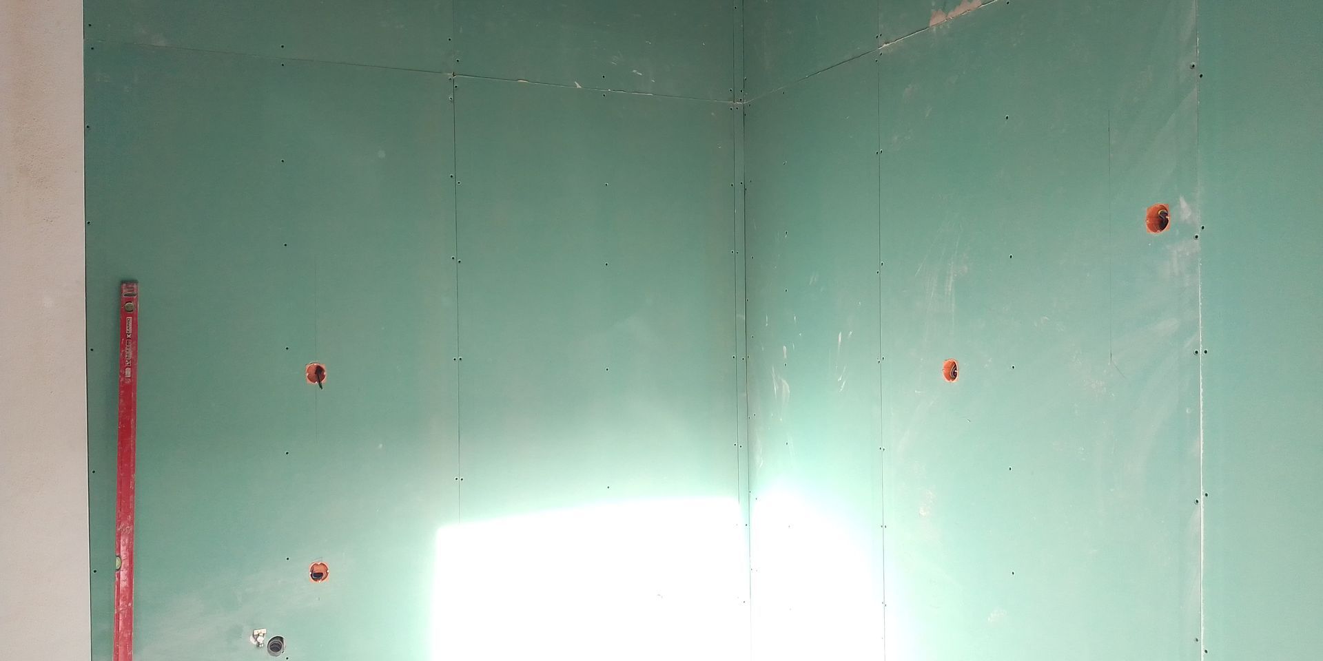 Canto de um cômodo com parede de gesso verde; vários buracos foram abertos na parede. Um cano vermelho é visível.