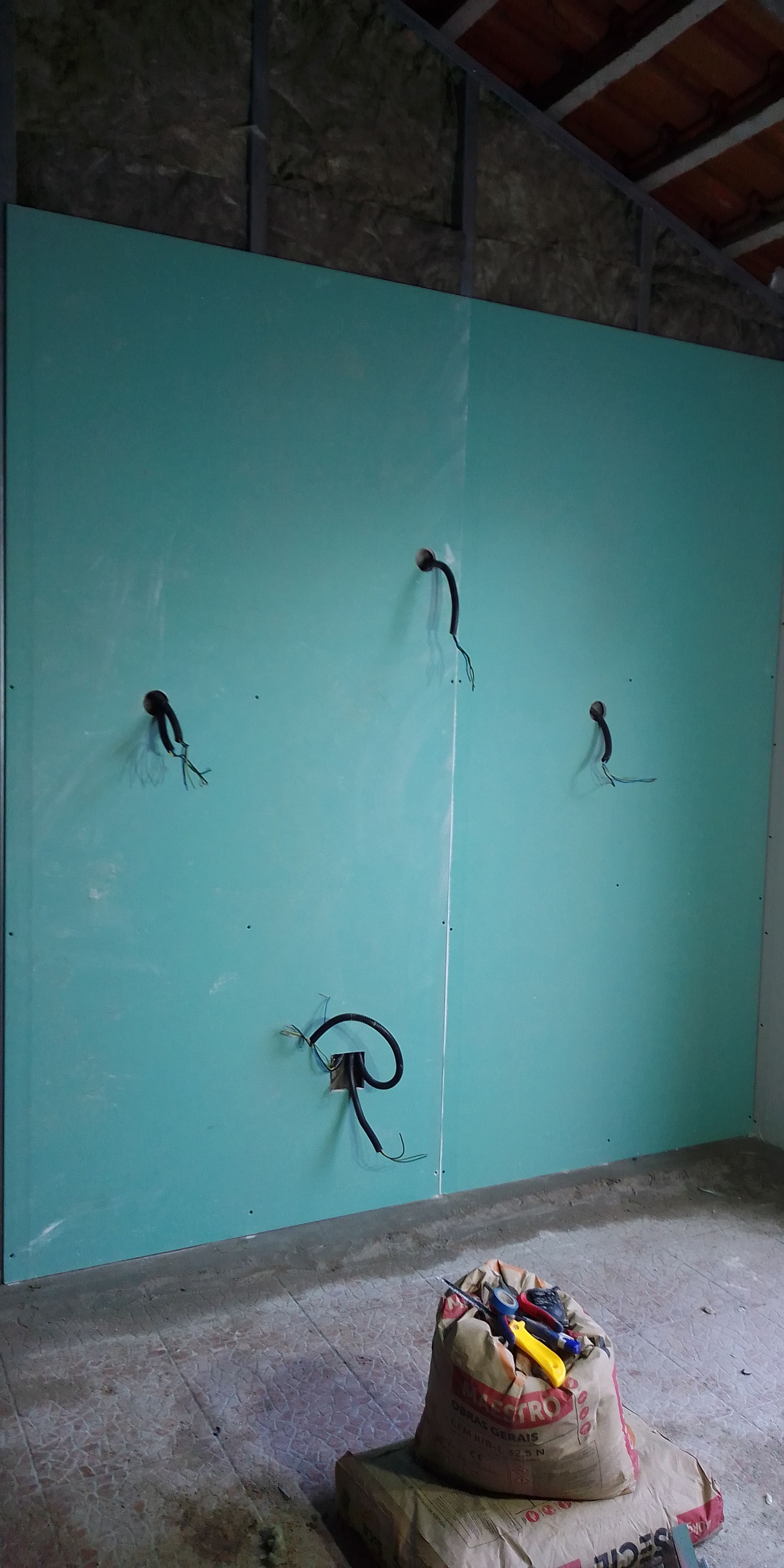 Parede azul-esverdeada com fiação elétrica em cômodo inacabado. Uma bolsa de ferramentas está no chão.