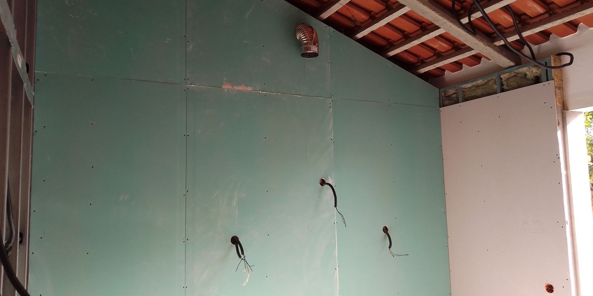 Interior de um cômodo com paredes de gesso acartonado verde. Fiação exposta e o início da construção do teto são visíveis.
