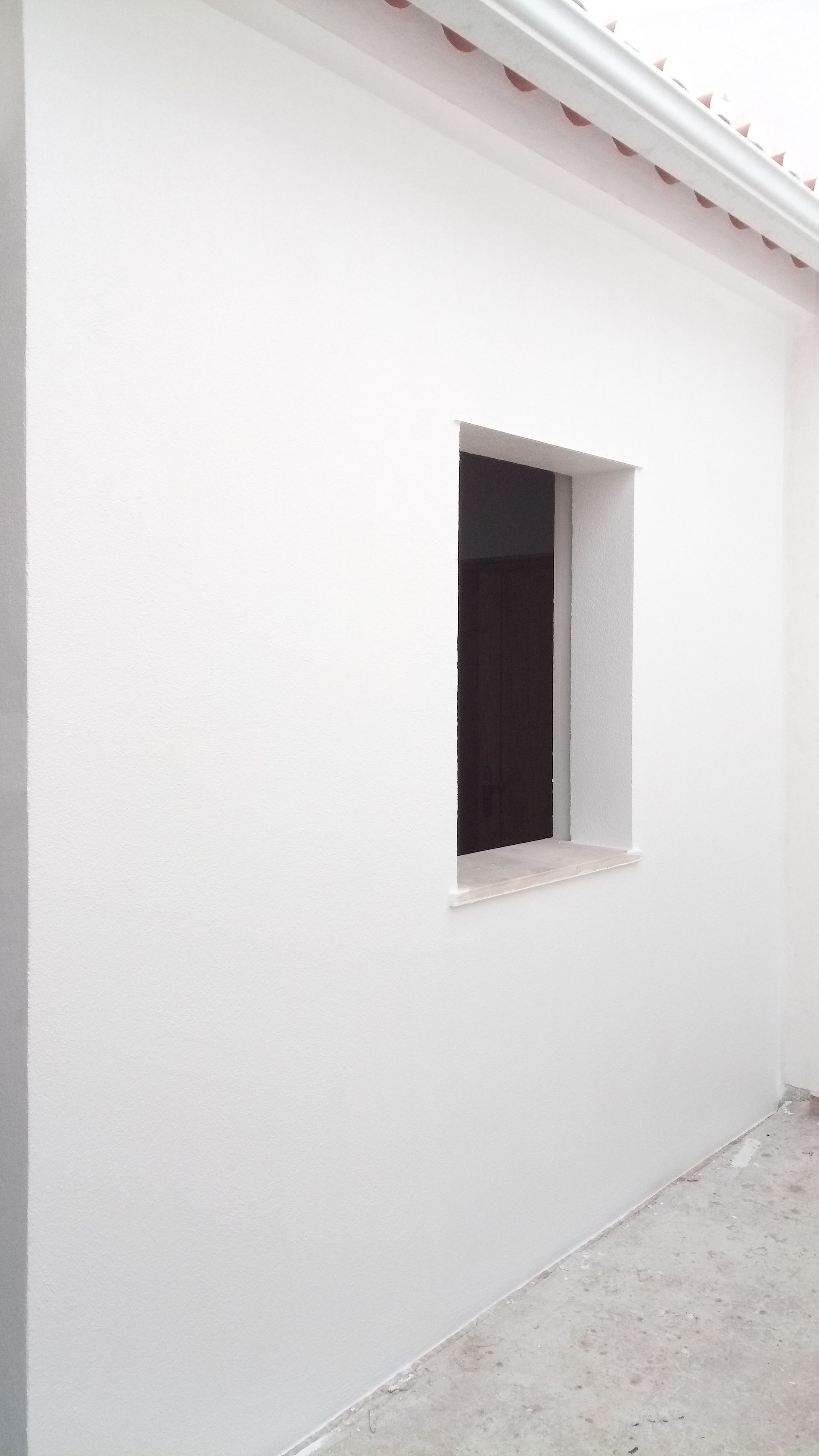 Parede externa branca com uma pequena janela, sob um telhado e calha.