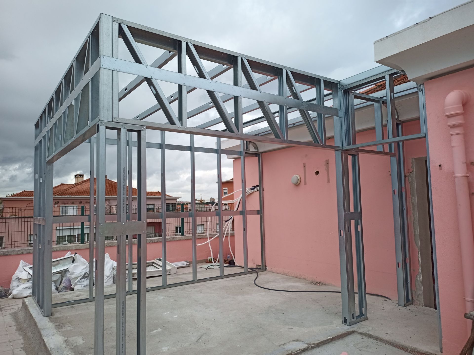 Estrutura metálica de uma construção em um telhado, com uma parede rosa e céu nublado ao fundo.