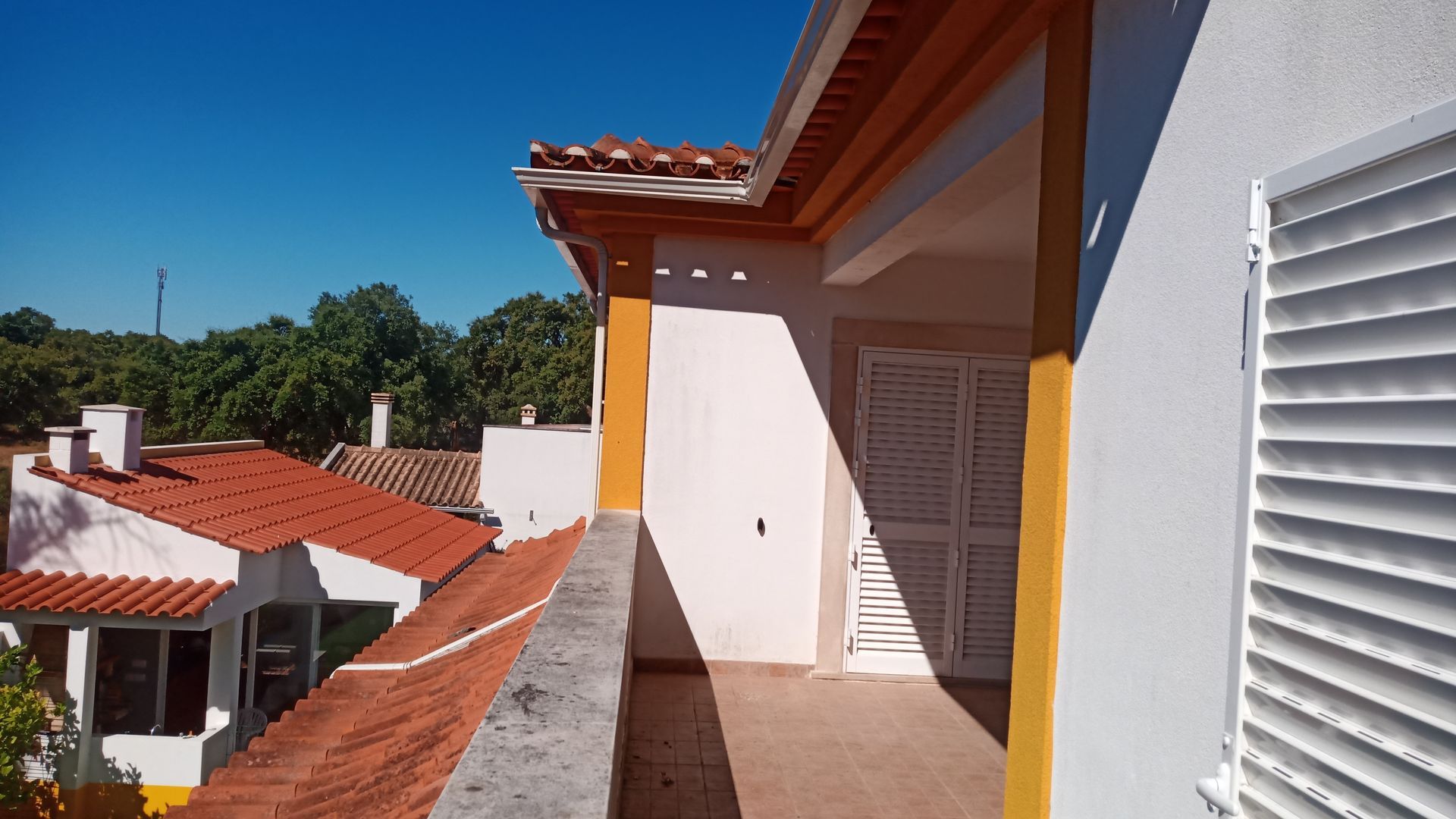Vista da varanda para prédios com telhados de telha vermelha, céu azul e venezianas brancas.
