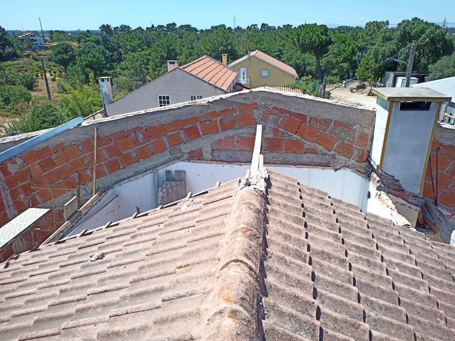 Terraço com telhas vermelhas, paredes de tijolos, chaminé e vista para casas e árvores sob um céu limpo.