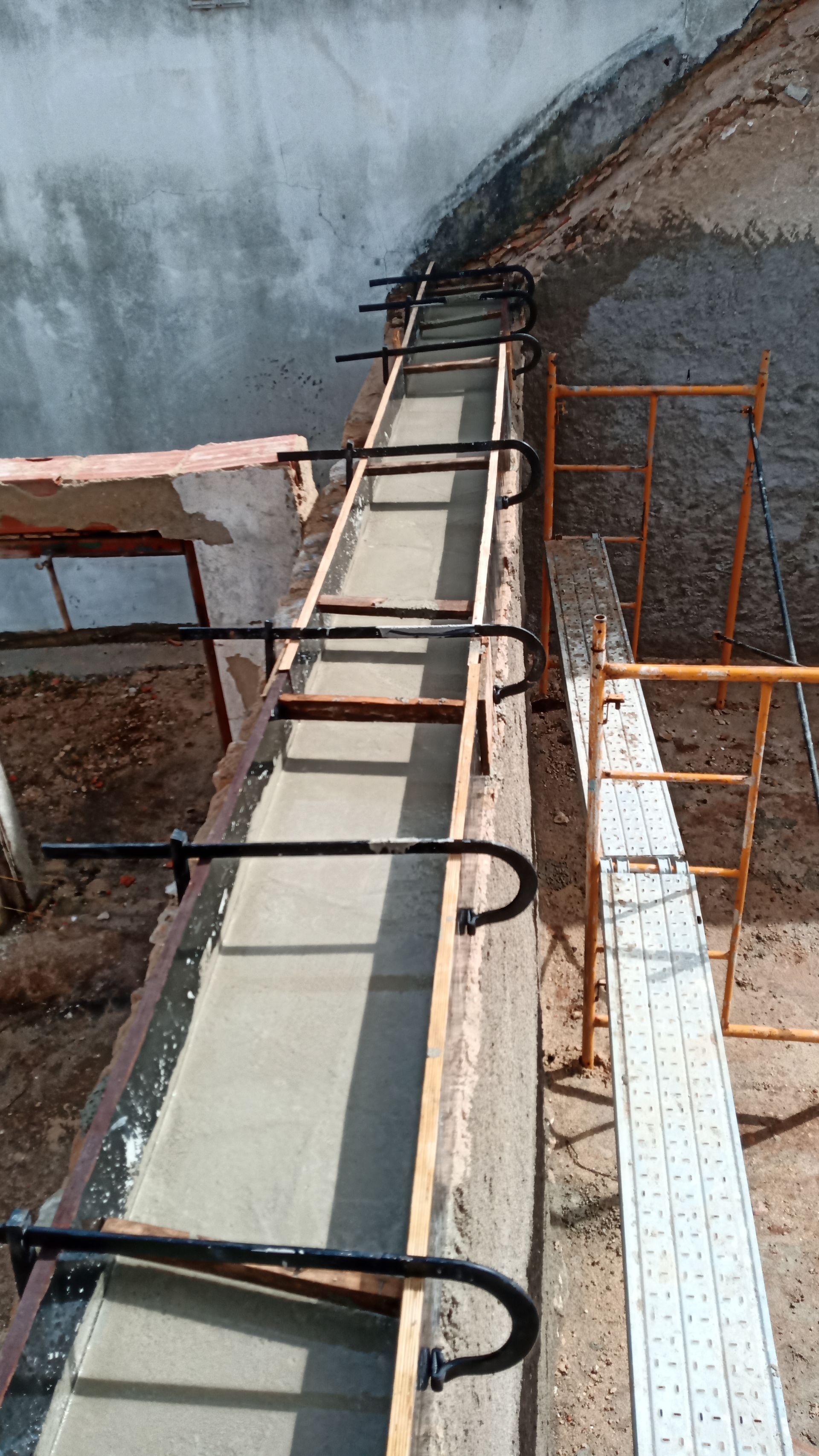 Concreto recém-moldado em uma forma longa e estreita, sustentado por suportes de metal preto; ambiente externo.