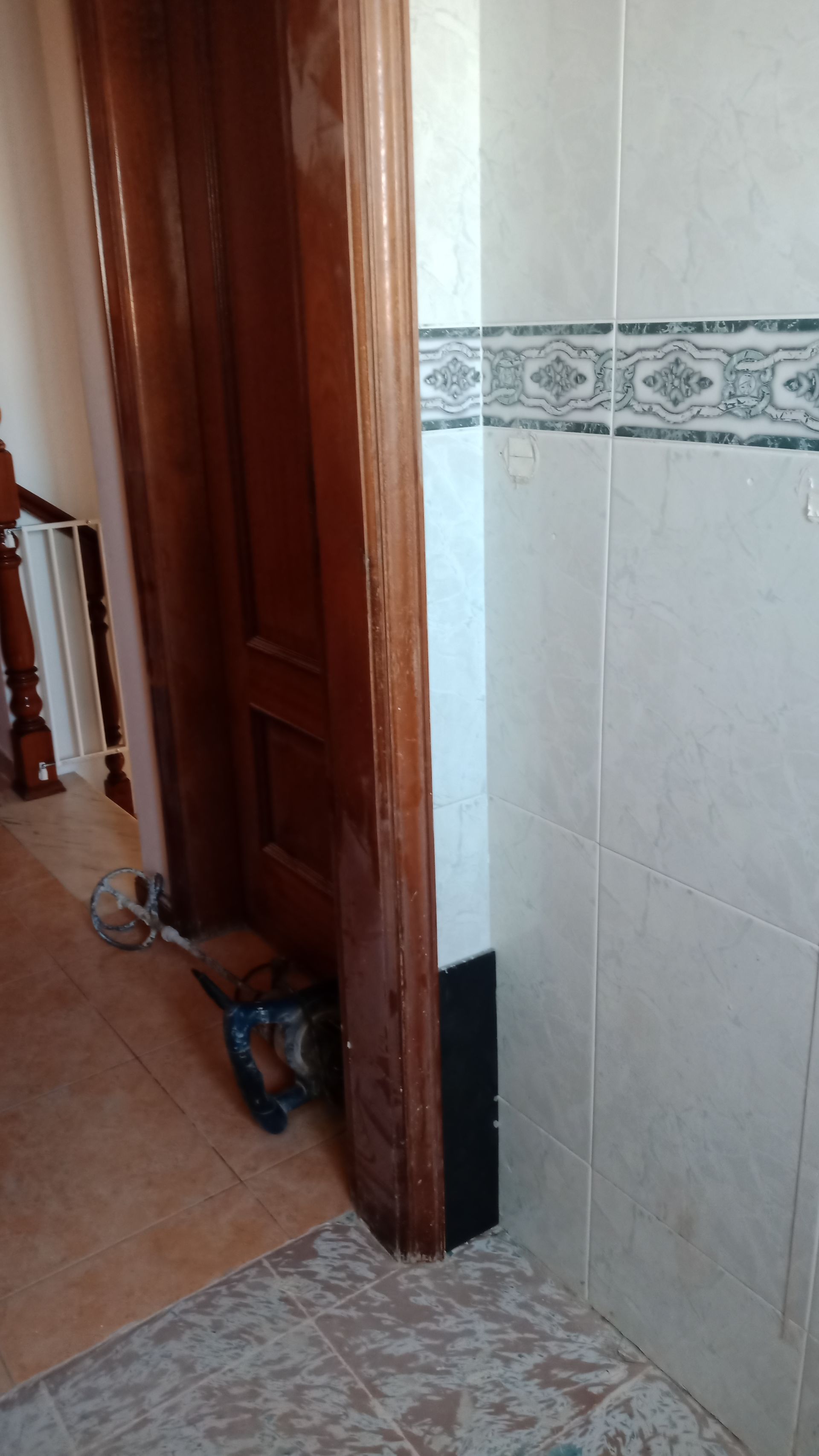 Batente de porta de madeira ao lado de uma parede revestida de azulejos. Um objeto preto é parcialmente visível sob a porta.