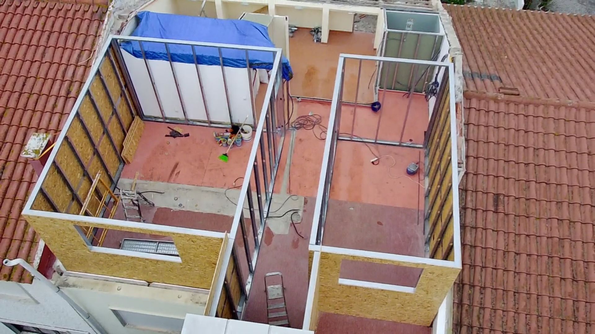 Canteiro de obras: estruturas parcialmente construídas com a estrutura exposta e uma lona azul no telhado, vista de cima.