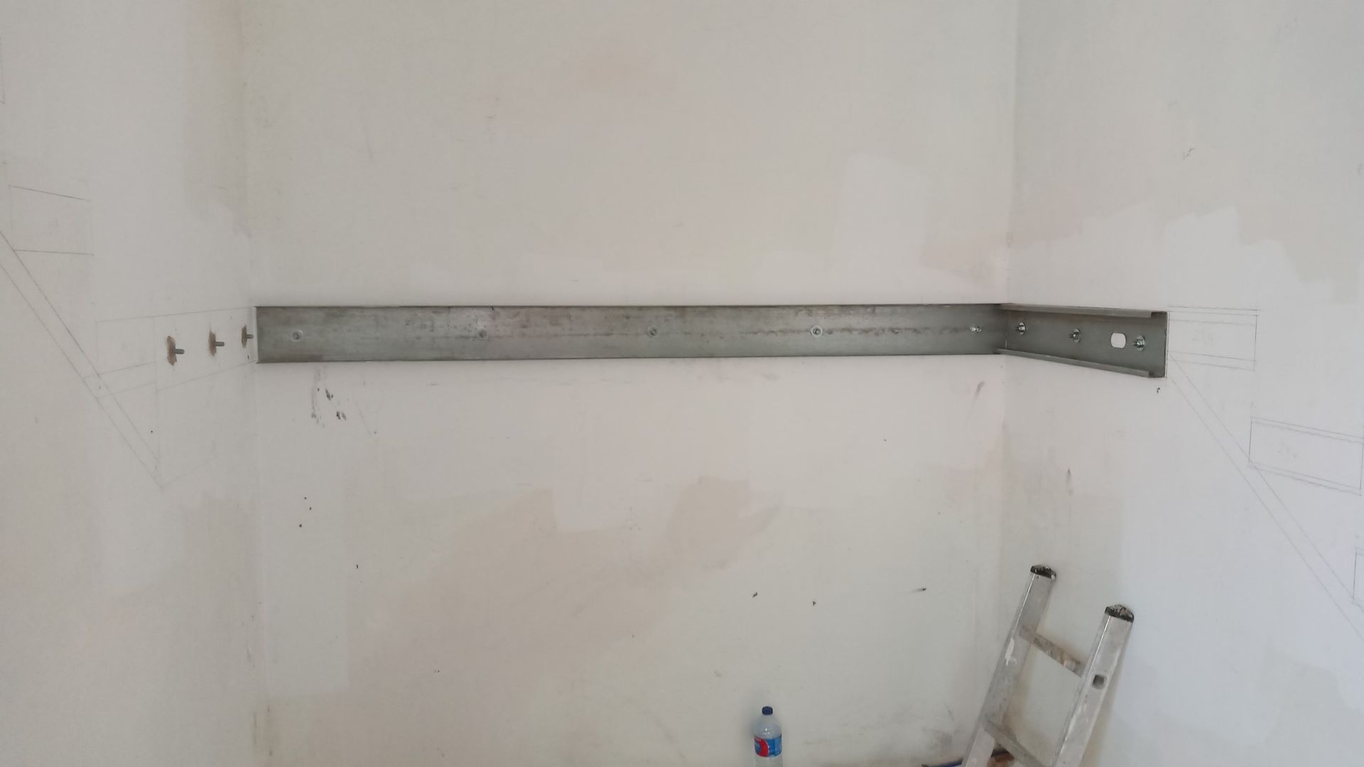 Suportes de prateleira de metal fixados em uma parede branca em um cômodo; uma escada está posicionada abaixo.