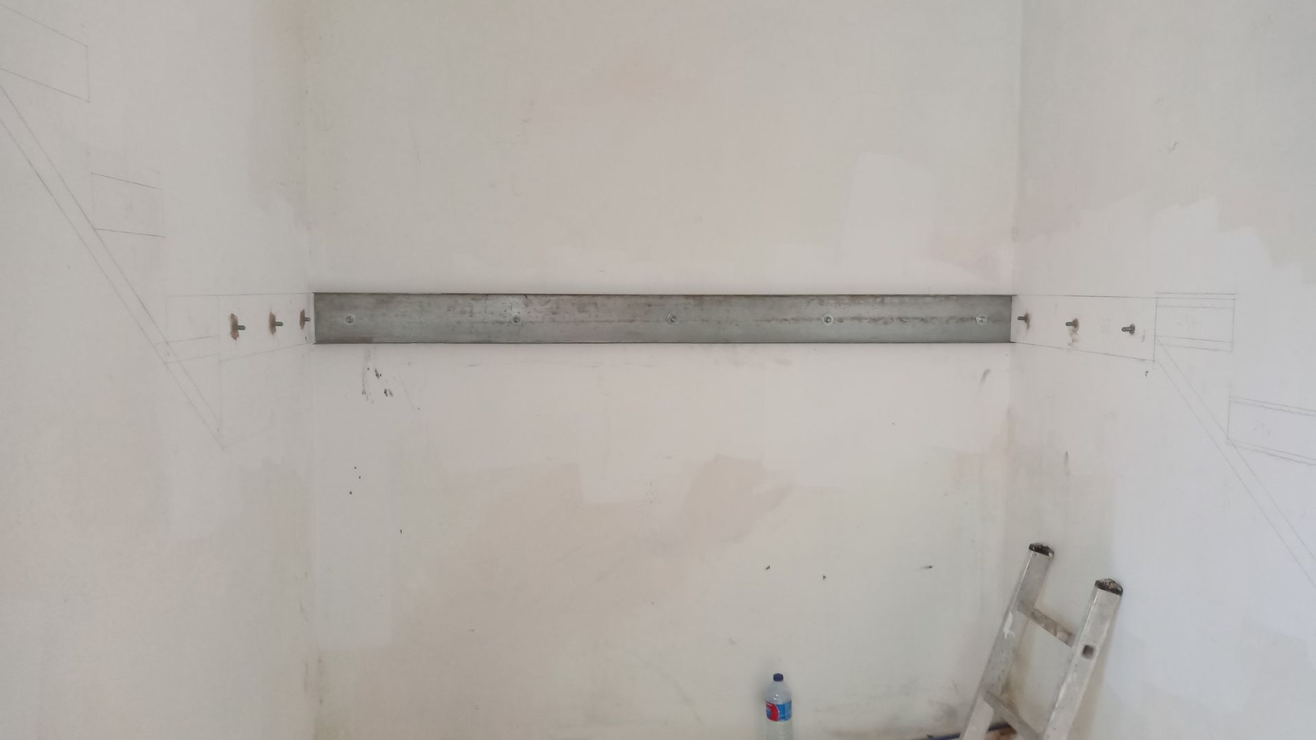 Viga de sustentação metálica fixada em uma parede branca em uma sala; uma escada está no chão.