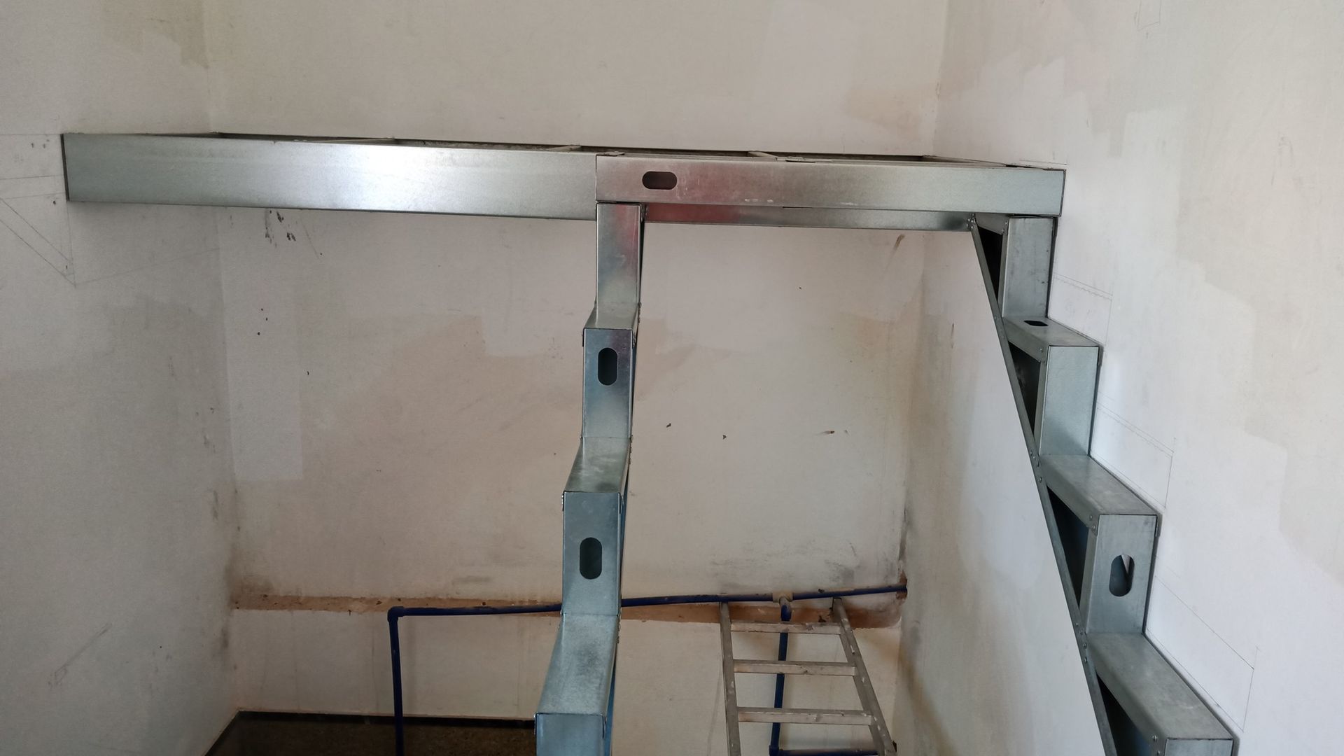 Estrutura metálica de escada que leva a uma pequena plataforma superior inacabada em uma sala com paredes brancas.
