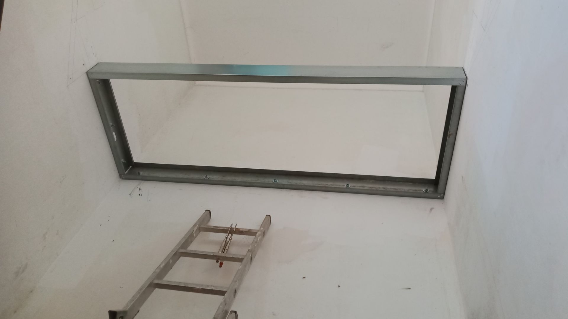 Estrutura metálica instalada em uma abertura na parede branca; uma escada está posicionada abaixo.