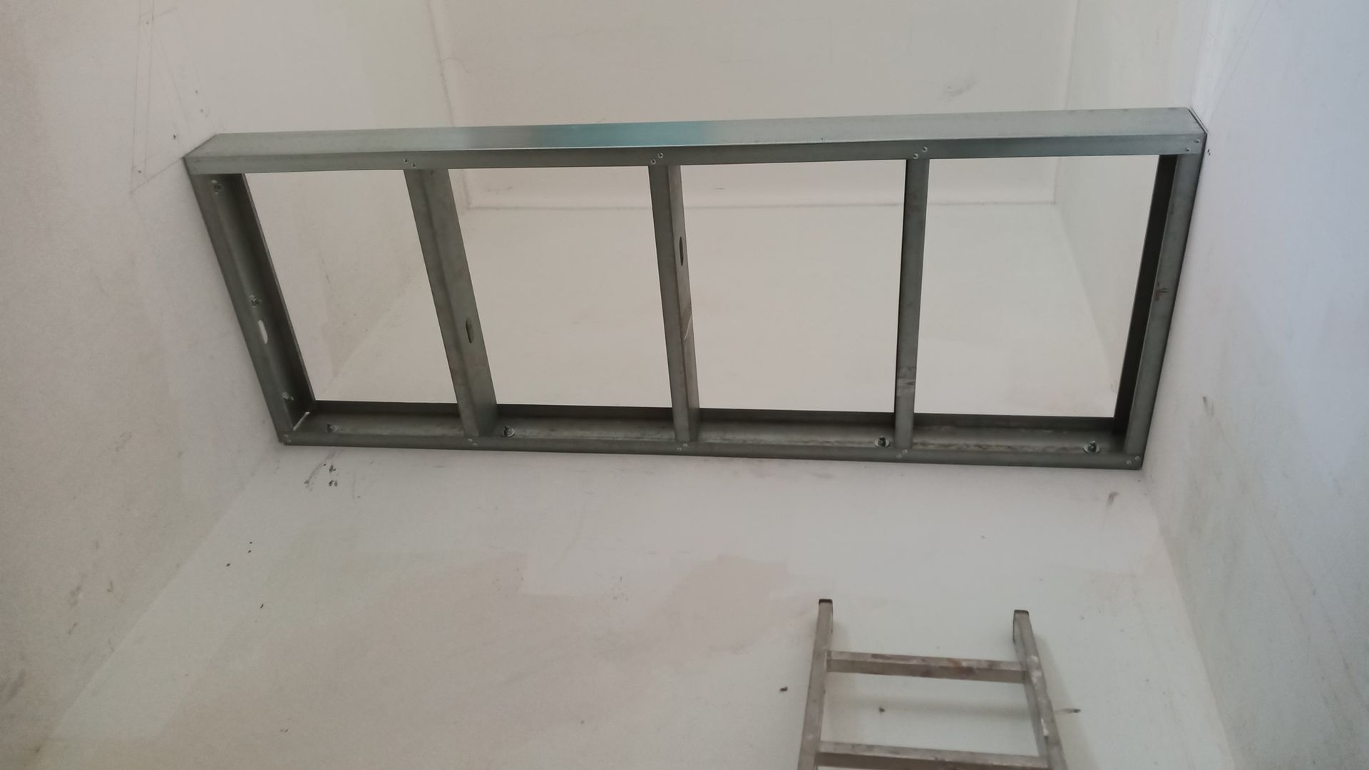 Estrutura metálica instalada no teto, provavelmente para uma luminária ou claraboia. Paredes brancas, pequena escada visível.
