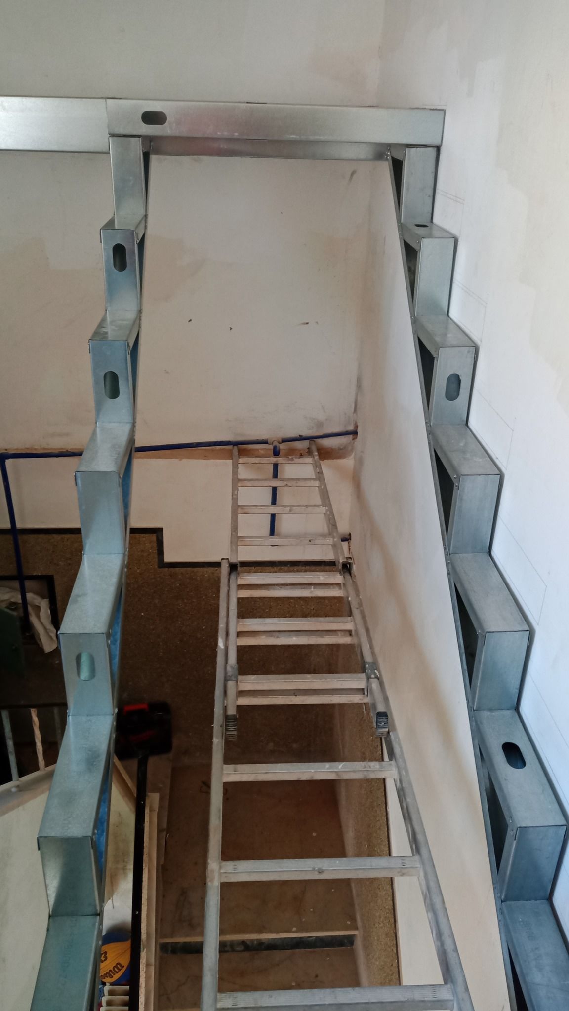 Escada metálica que leva a um andar superior, com uma escada de mão à frente, ao longo de uma parede branca.