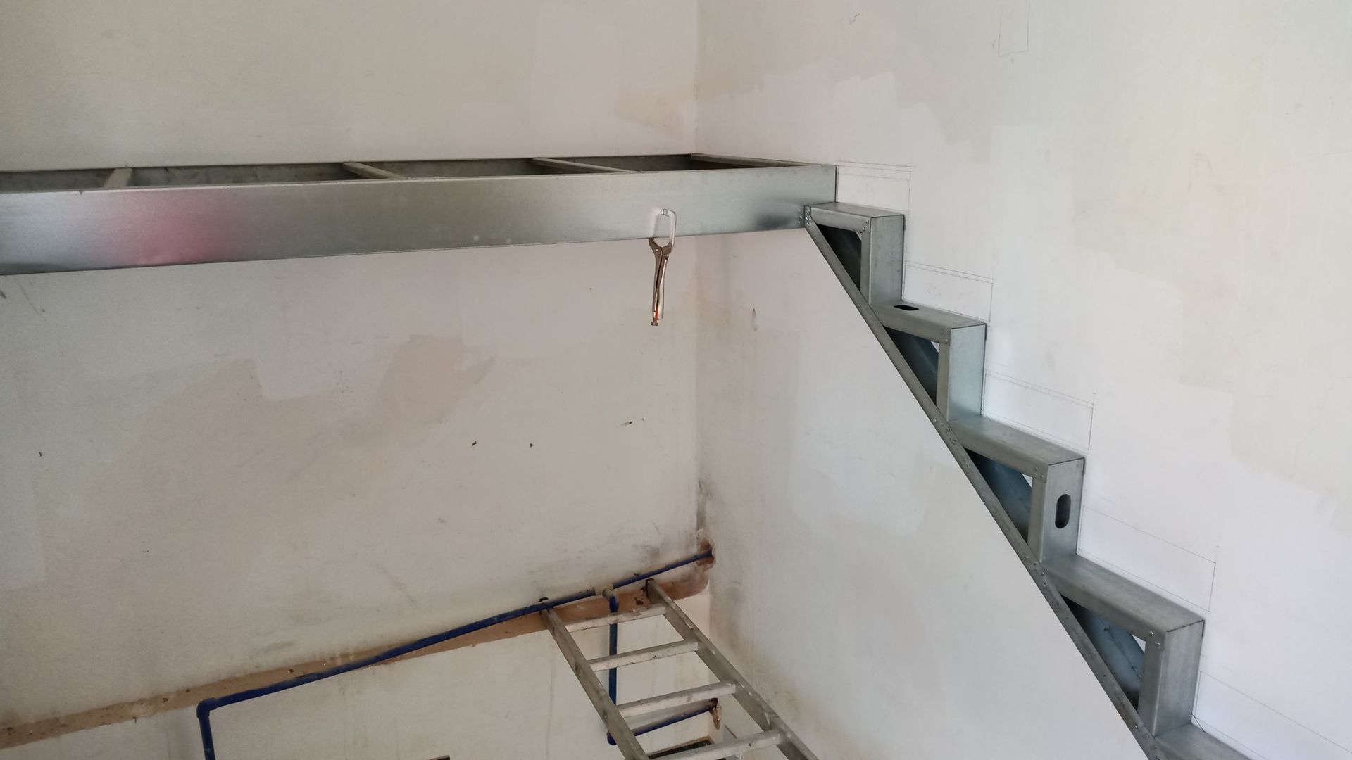 Construção de escada com estrutura metálica. Degraus e estrutura da plataforma superior visíveis, paredes de gesso cartonado sem acabamento.