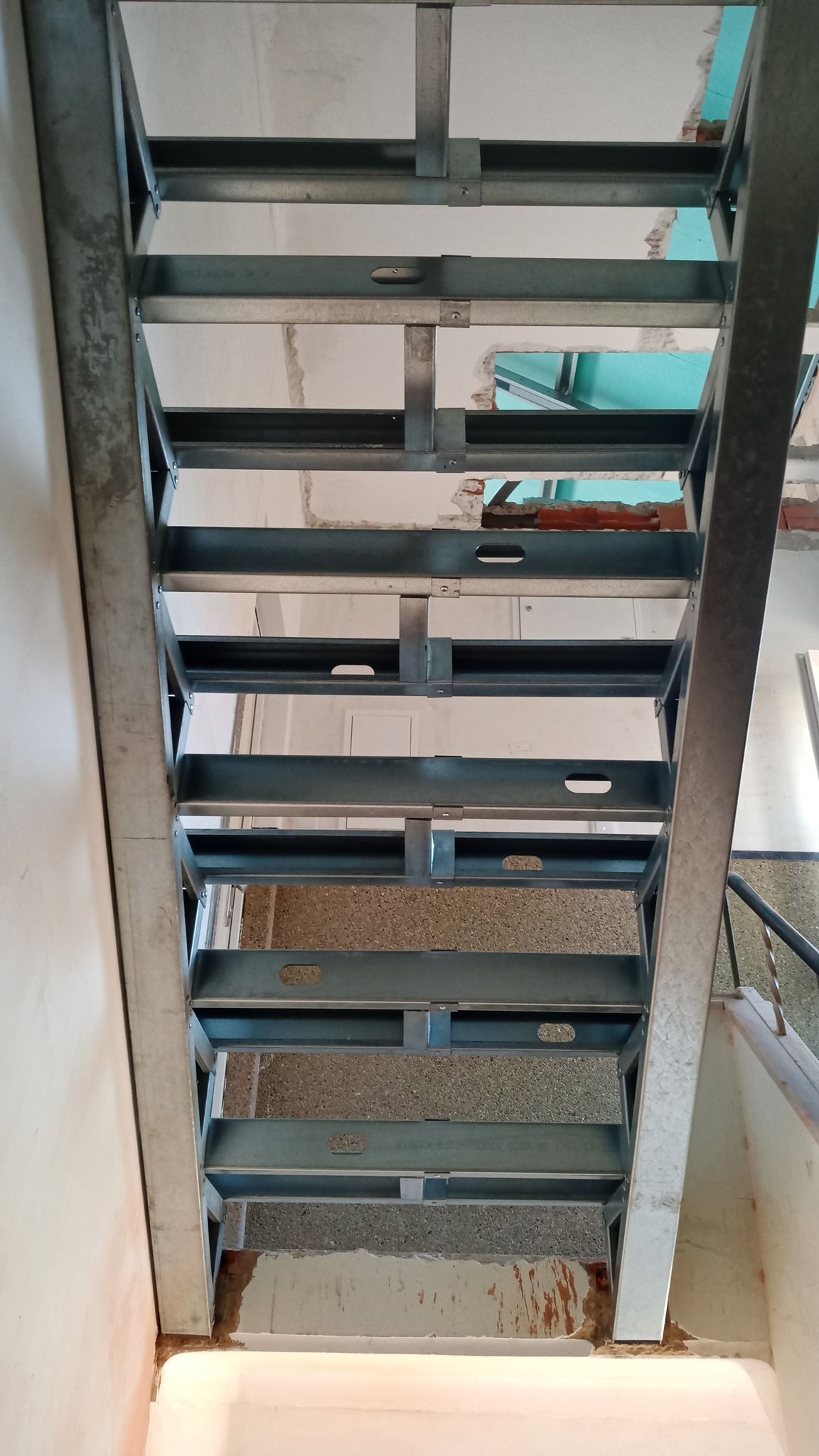 Estrutura metálica de escada com degraus, vista de baixo. Metal cinza em contraste com uma parede de cor clara.