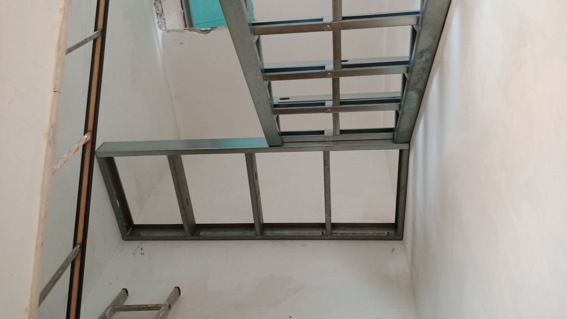Estrutura metálica para teto rebaixado em uma sala com paredes brancas; uma escada é visível.