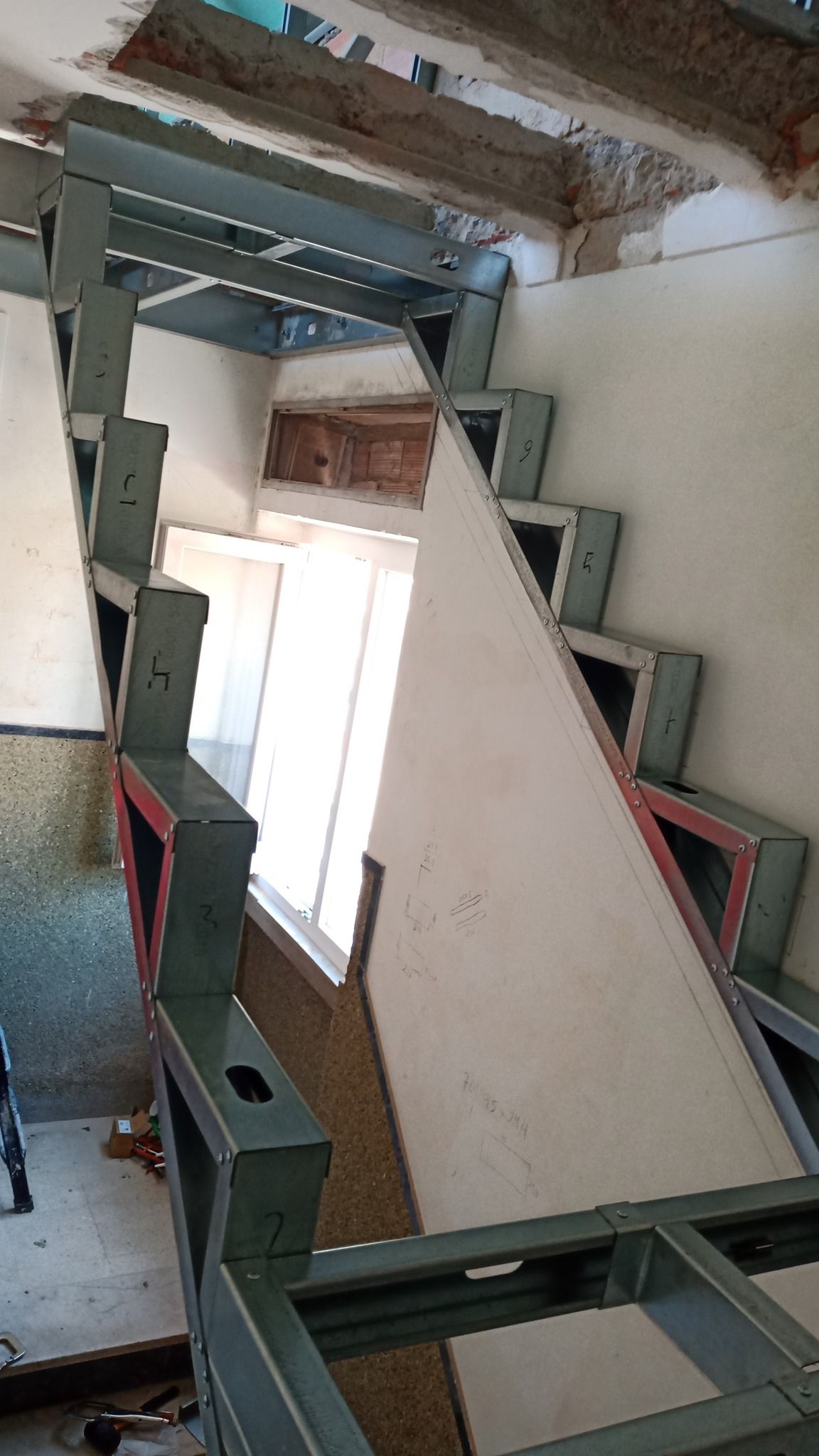 Escada com estrutura de aço em construção, com espelhos abertos e estrutura de suporte visível, próxima a uma janela.