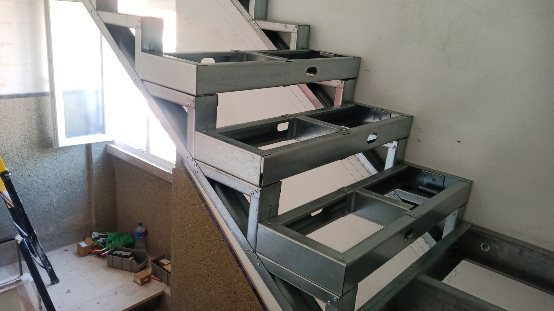 Estrutura metálica de uma escada inacabada encostada na parede, perto de uma janela.