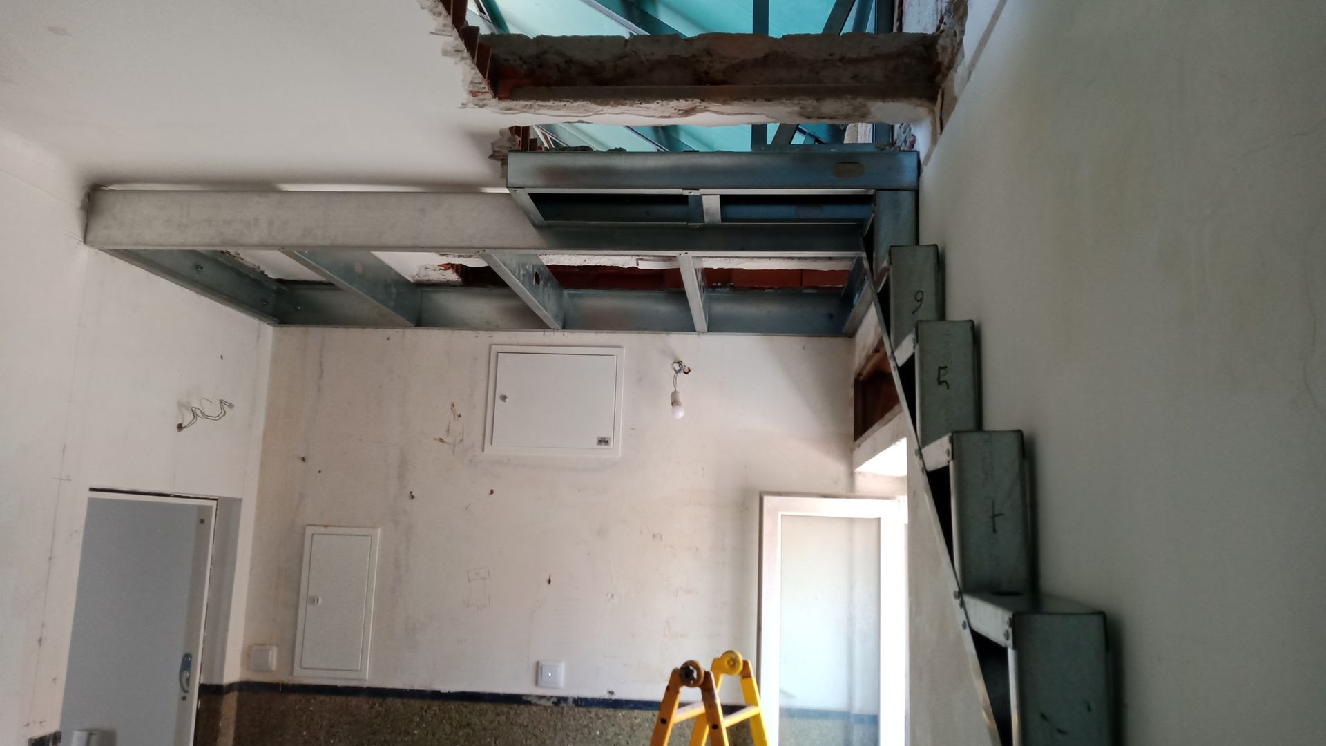 Interior em construção; escada com estrutura metálica e teto que levam a uma abertura.