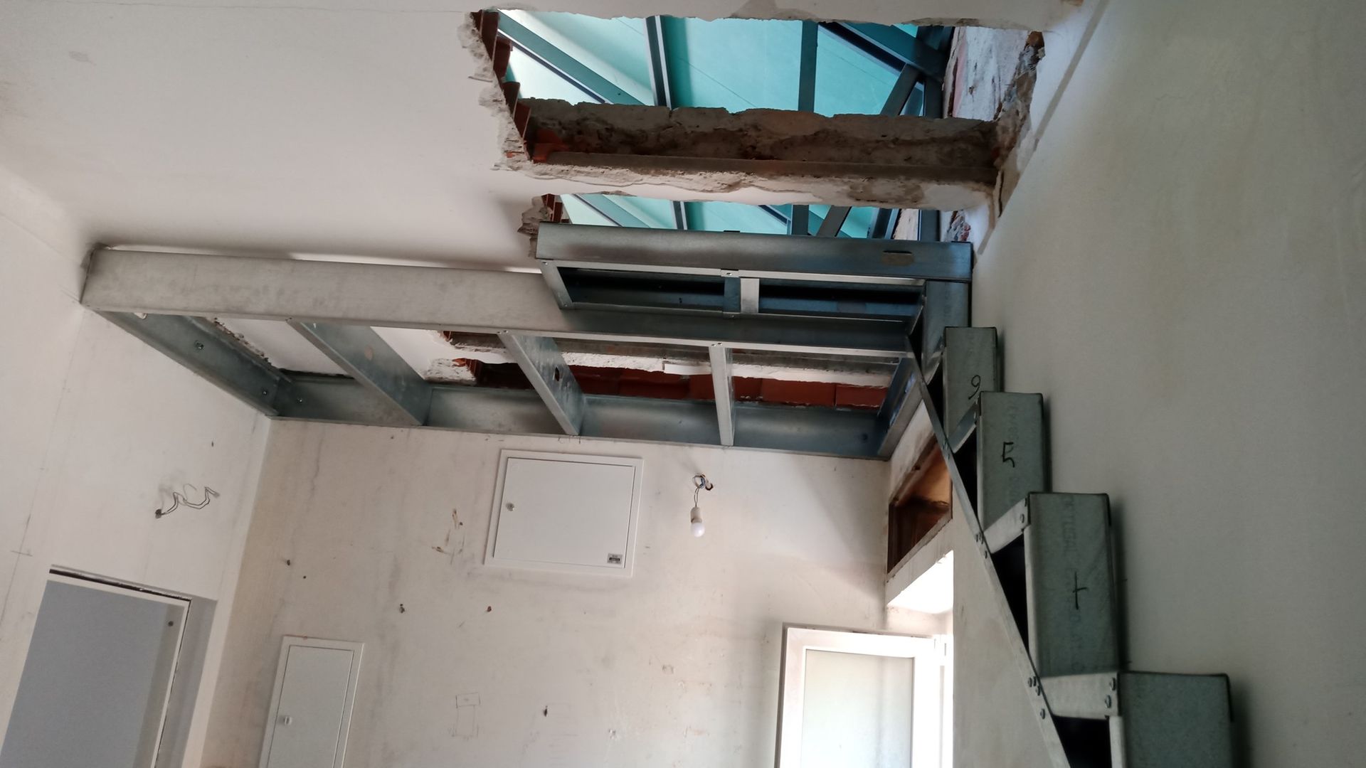 Vista interna de um canteiro de obras com uma escada metálica parcialmente construída que leva a uma plataforma próxima a uma abertura no teto.