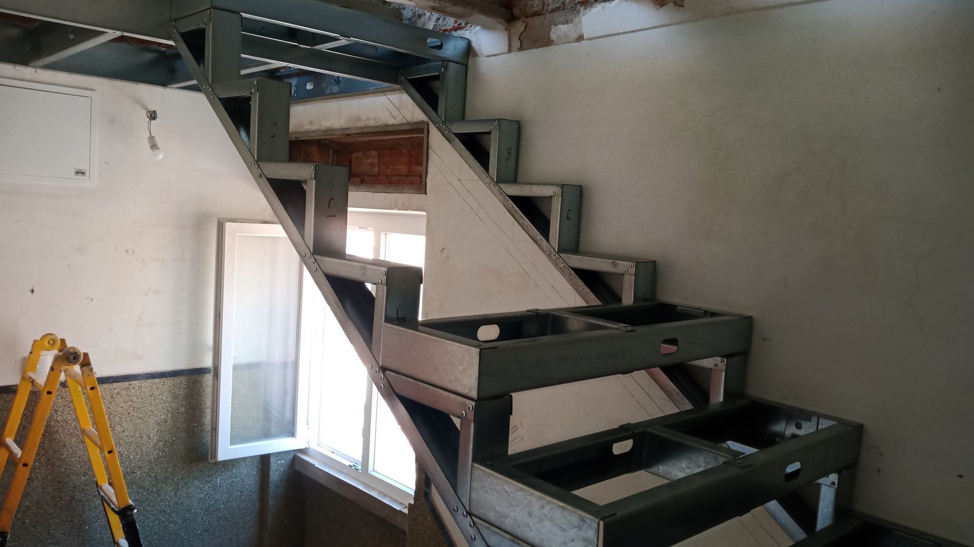 Escada em construção, com estrutura de madeira aparente, que dá acesso ao piso superior. Vista interior, ao lado de uma janela.