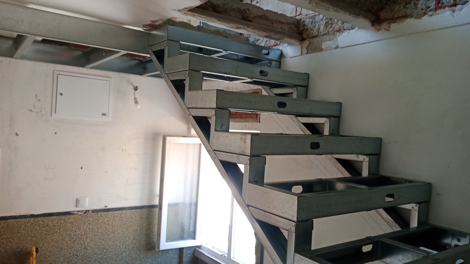 Escada metálica em um cômodo com paredes e teto nus, que leva ao andar superior. Janela com moldura branca.