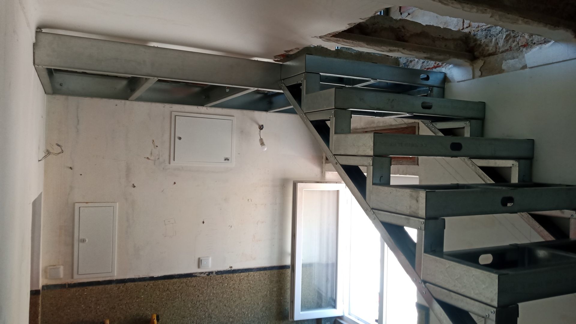 Escada metálica em um cômodo com teto danificado. Há uma janela no canto inferior direito e uma porta de serviço branca na parede.